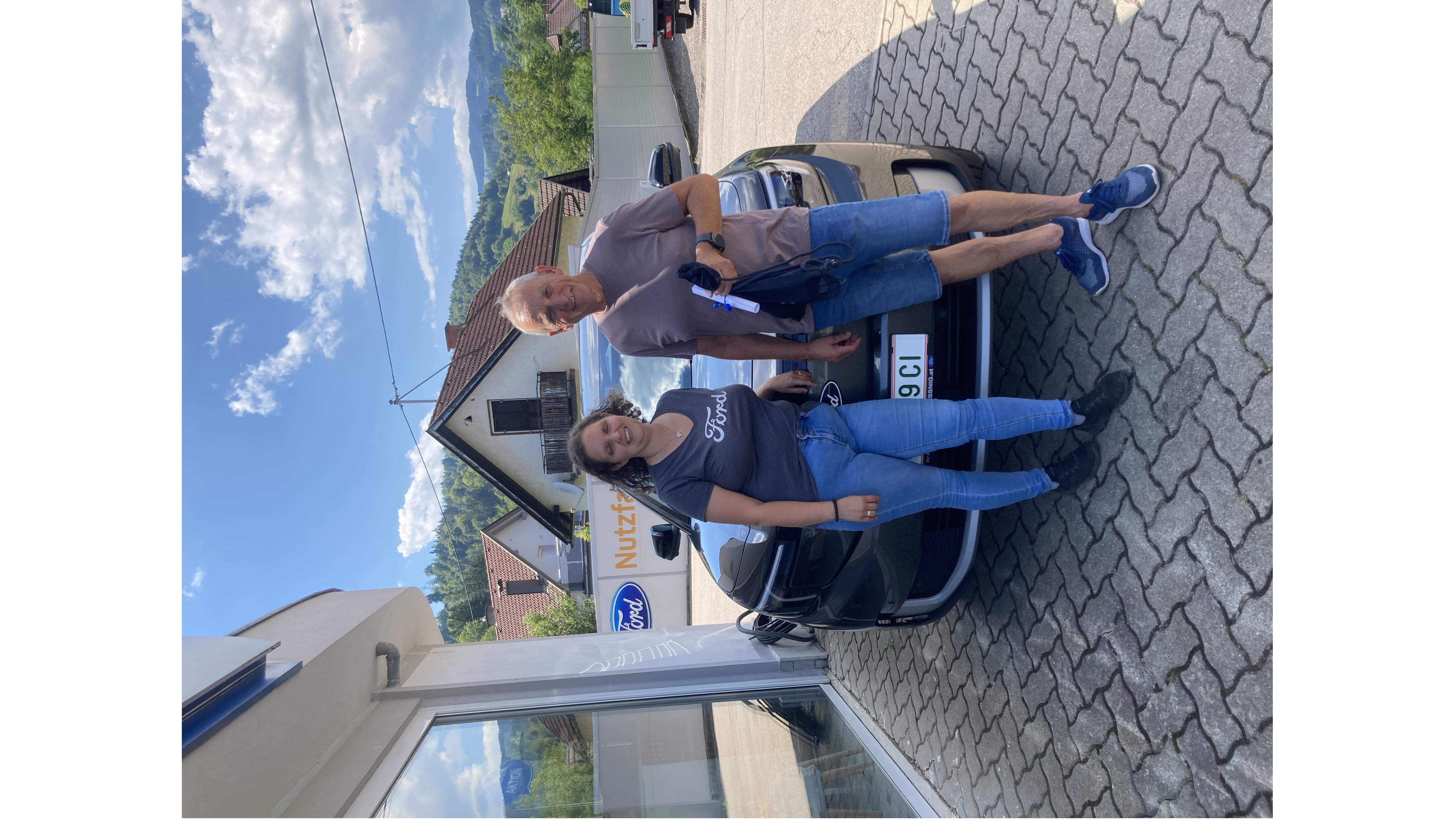 Zwei Personen posieren vor einem Ford-Auto bei einem Autohändler, umgeben von einer malerischen Landschaft.