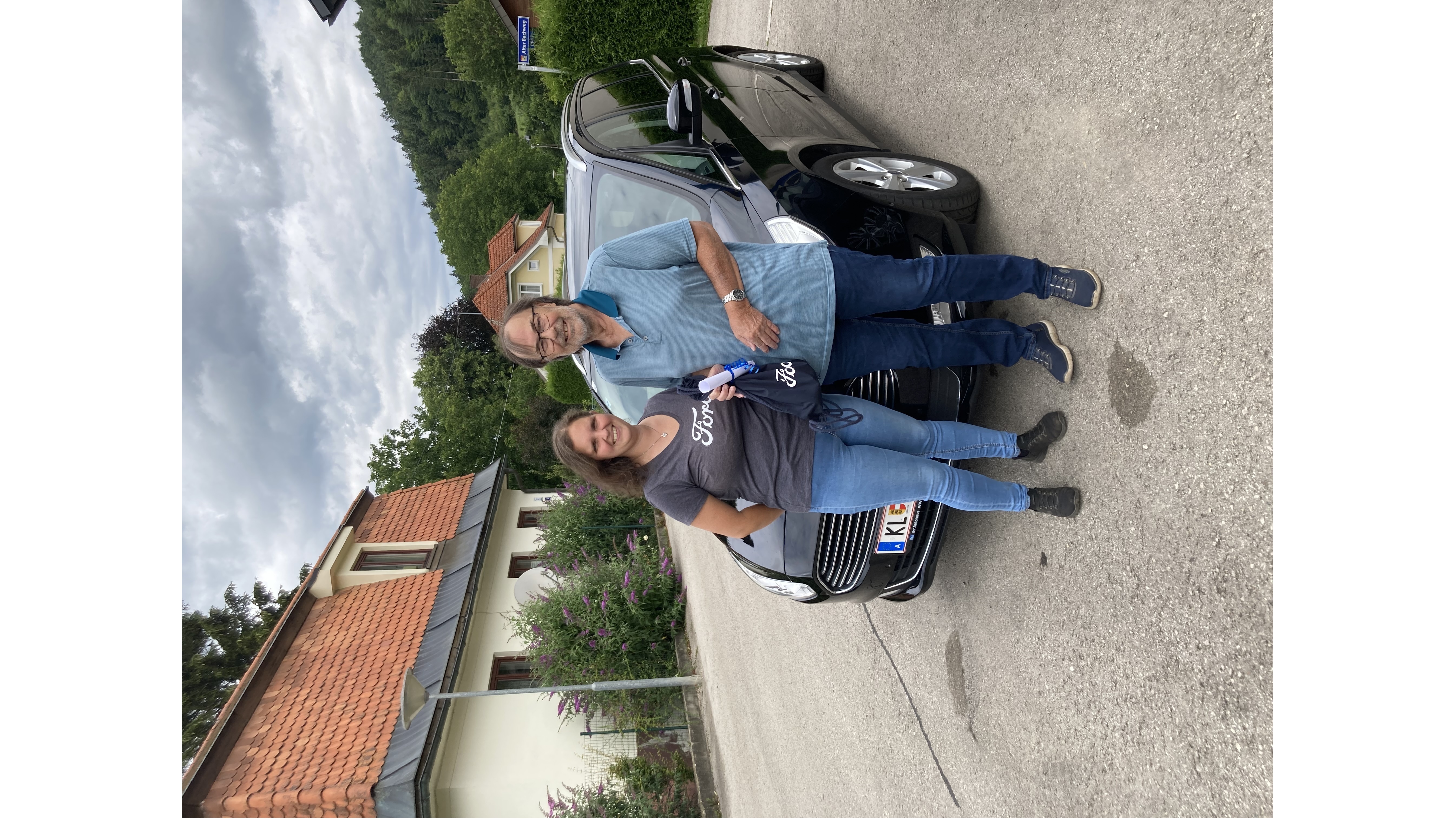 Eine Frau und ein Mann stehen neben einem schwarzen Auto auf einer Straße, umgeben von Bäumen und Häusern.