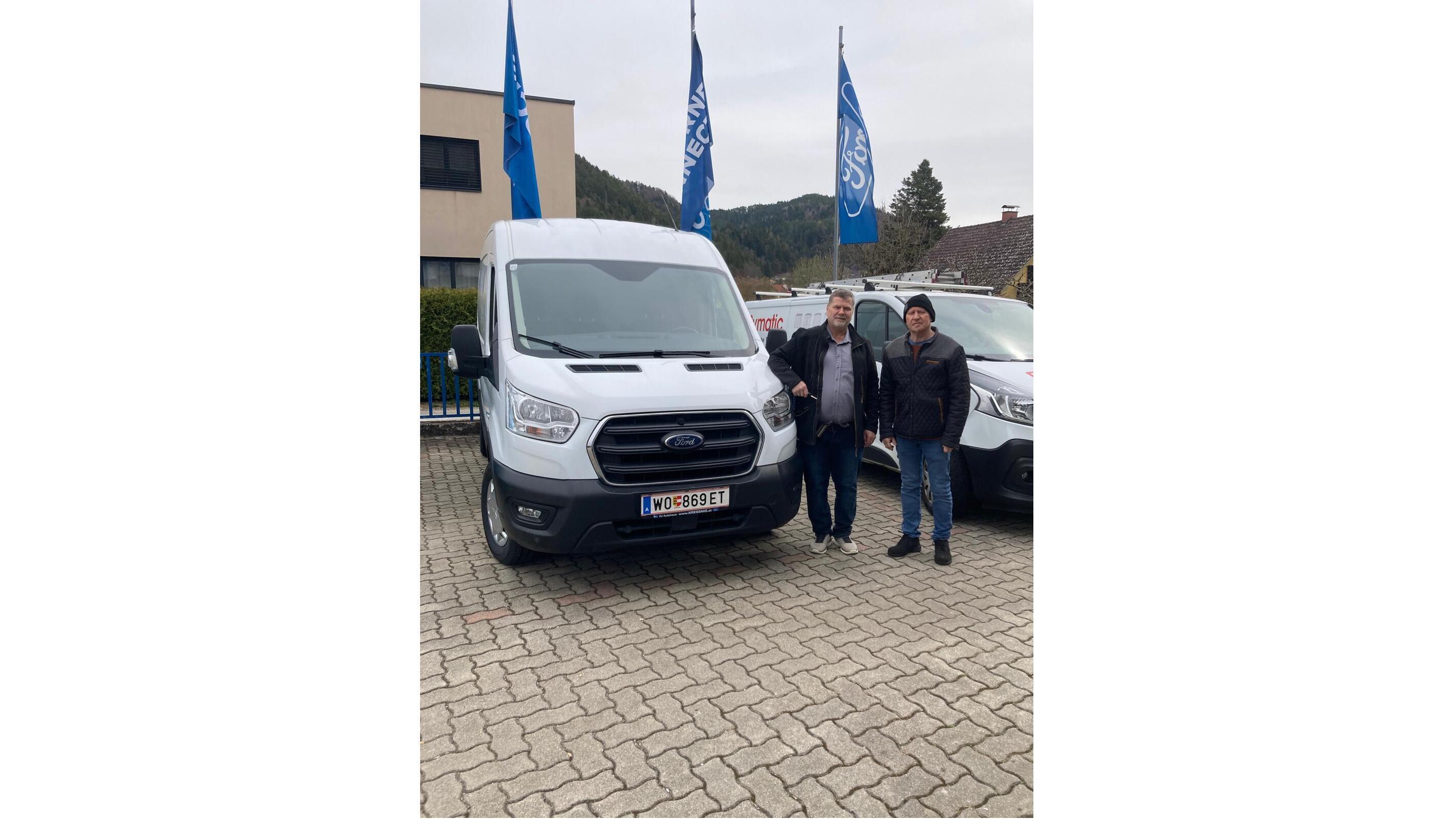 Zwei Männer stehen neben einem Ford Transporter auf einem Parkplatz, umgeben von Flaggen und Bergen im Hintergrund.