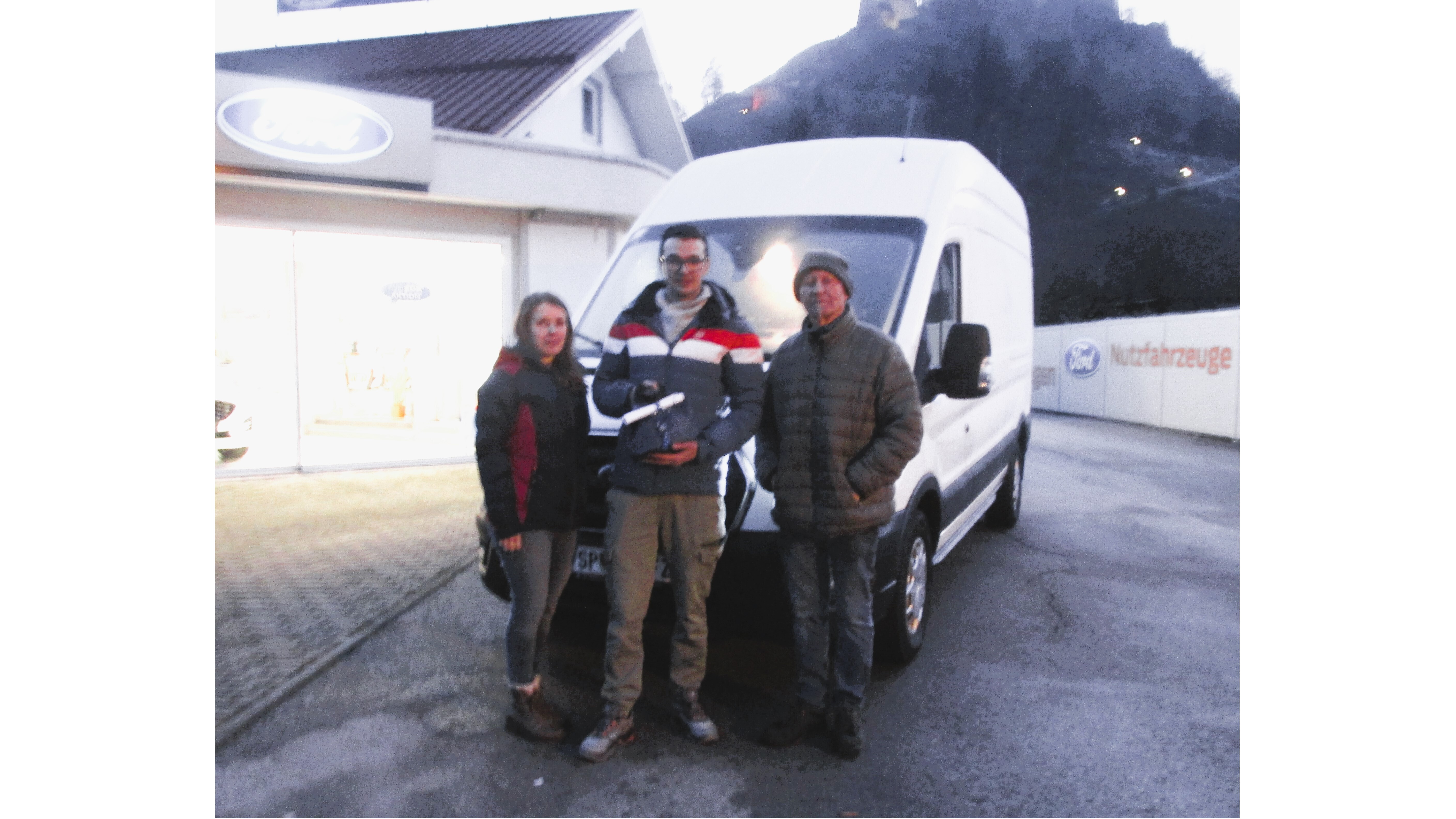 Drei Personen stehen vor einem weißen Lieferwagen bei einem Autoloadcenter. Im Hintergrund ist ein Berg sichtbar.
