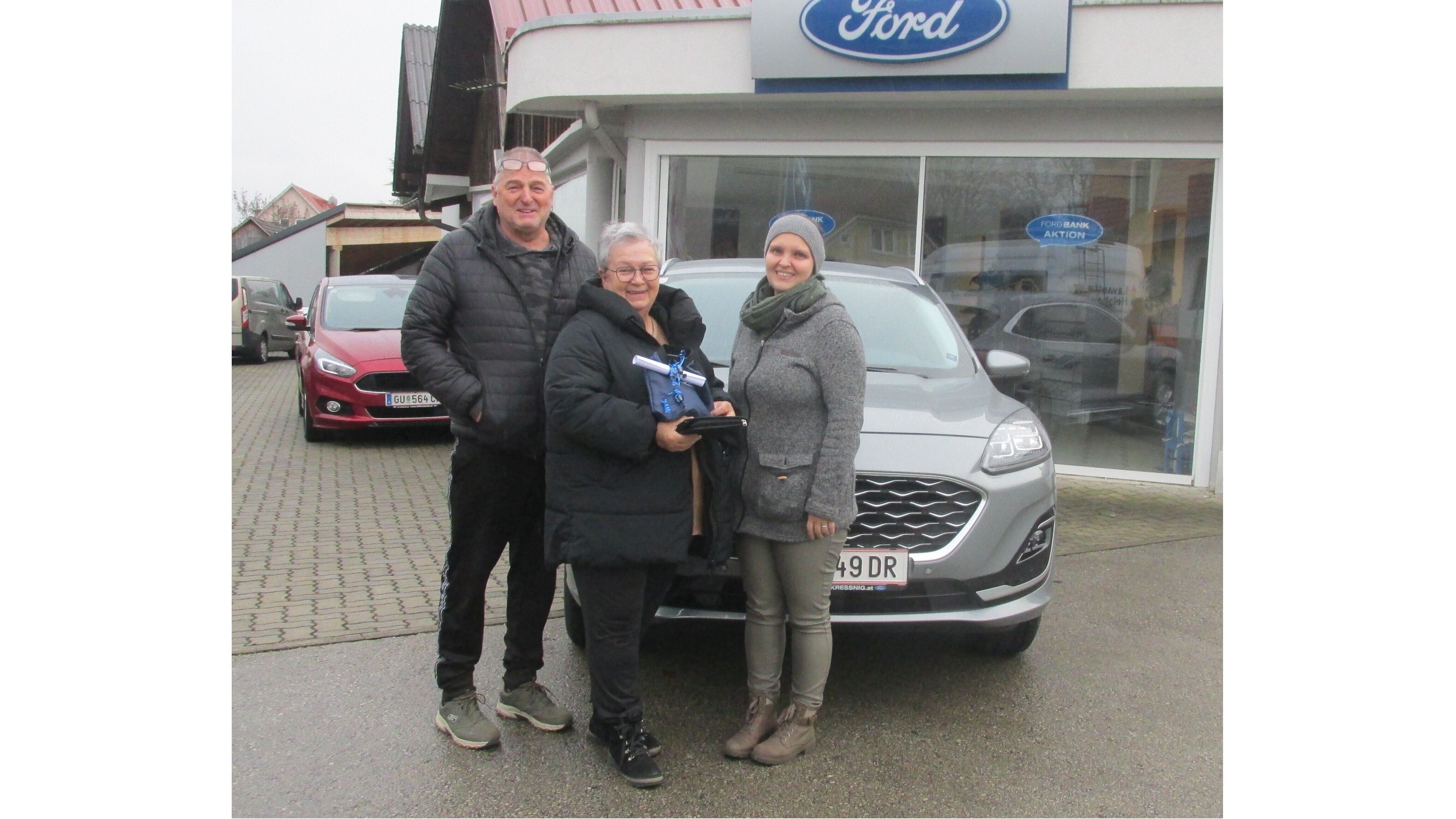 Drei Personen stehen vor einem Ford-Autohaus, lächelnd und mit einem Geschenk in den Händen. September 2023.