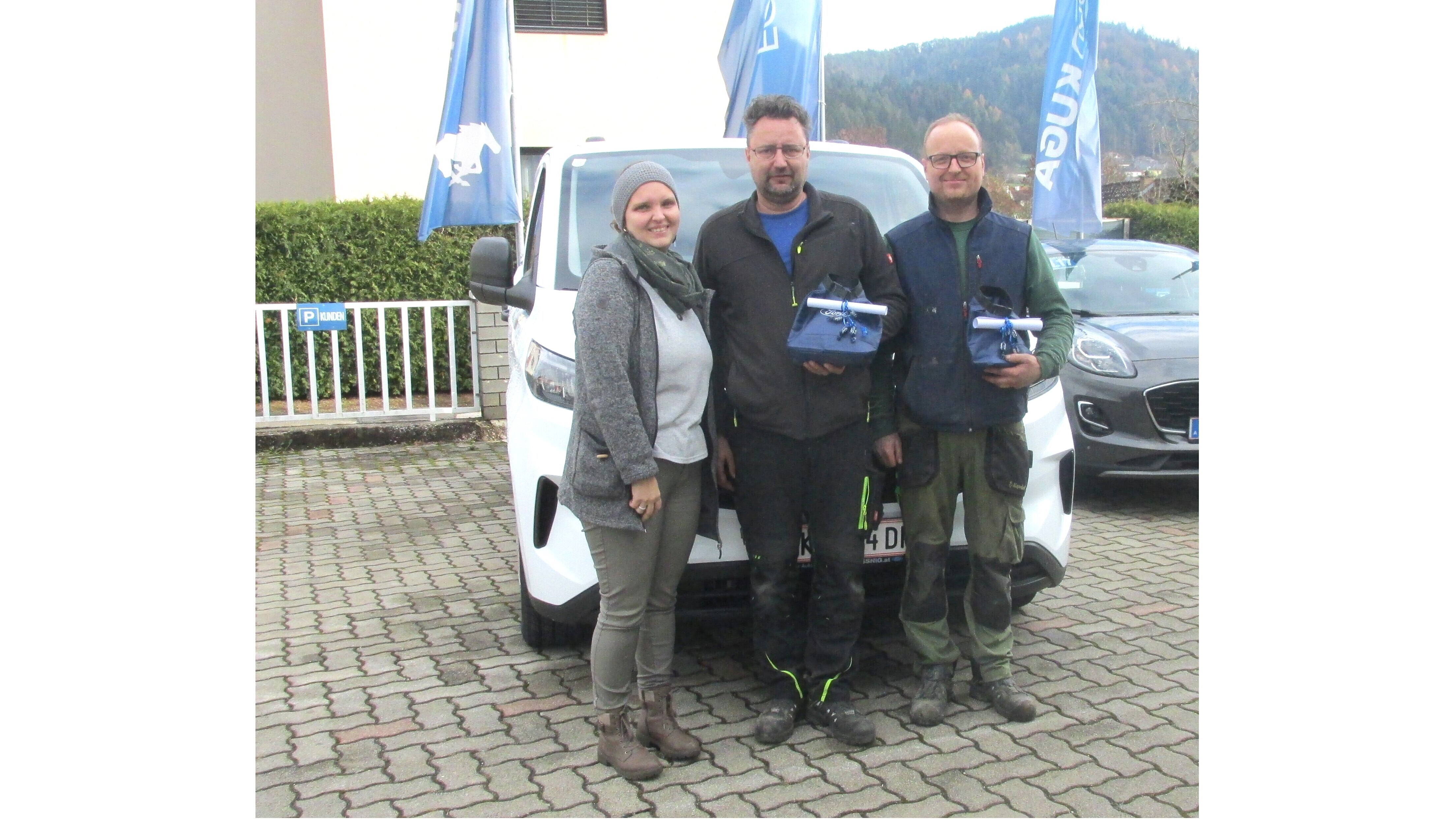 Drei Personen stehen vor einem weißen Fahrzeug, halten Geschenkpakete und posieren vor blauem Banner im Hintergrund.
