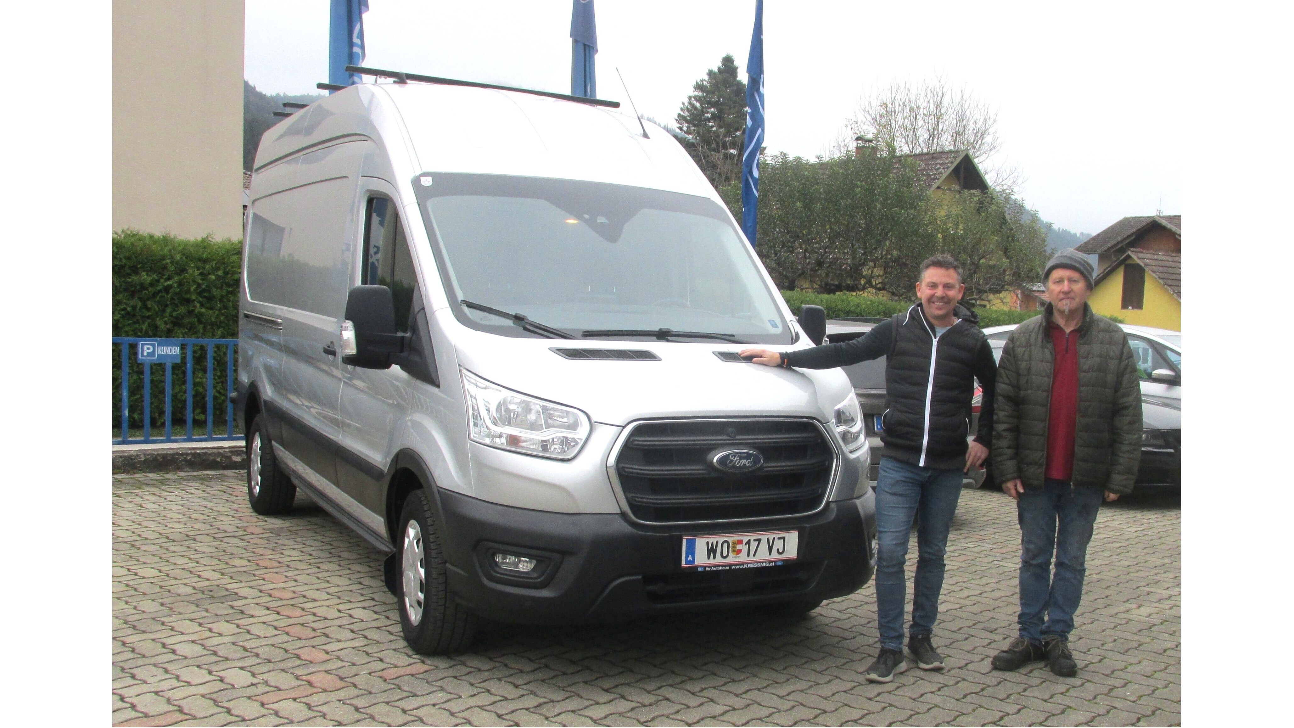 Zwei Männer stehen neben einem silbernen Ford Transporter auf einem Parkplatz, umgeben von grüner Landschaft.