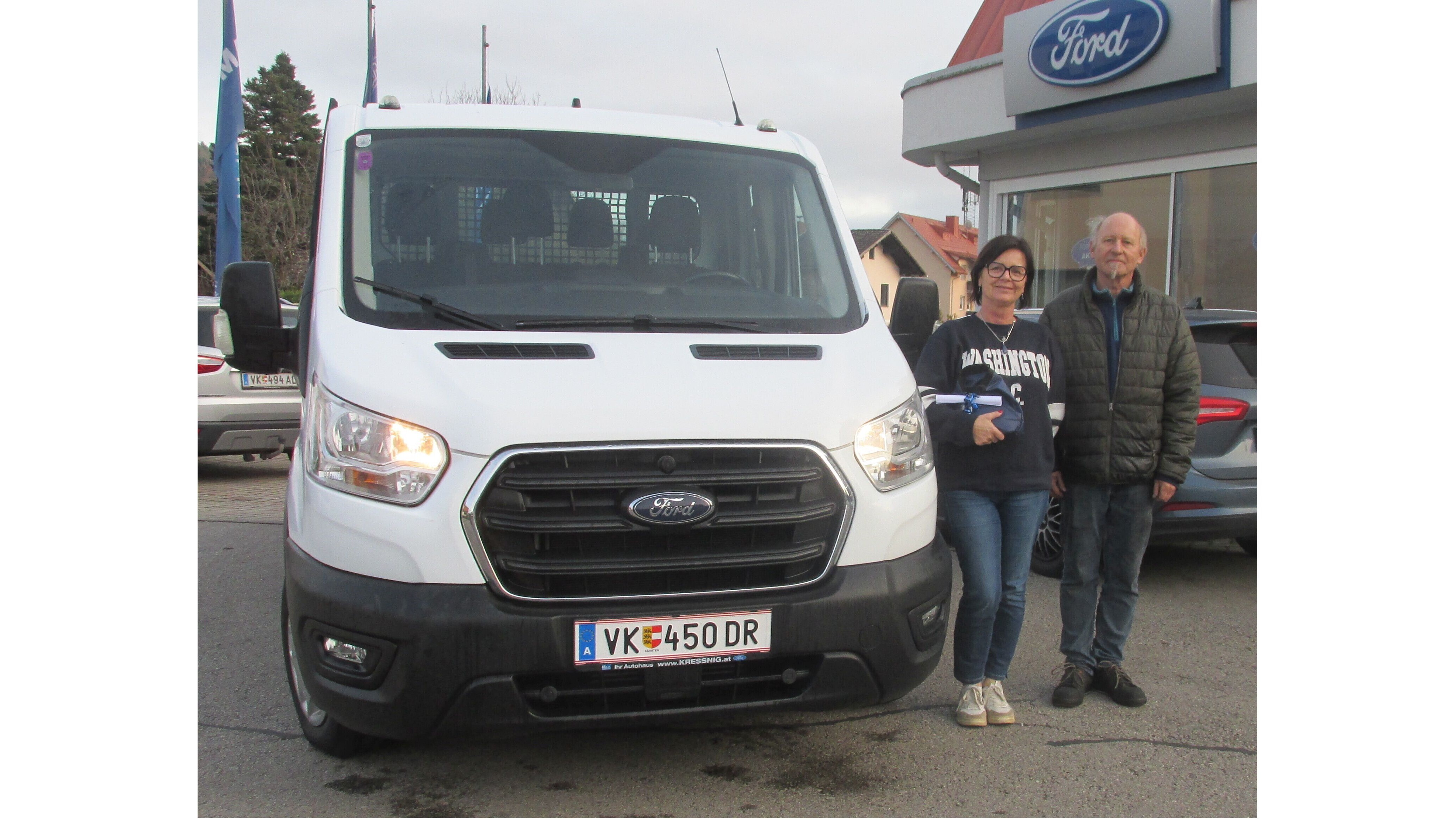 Eine Frau und ein Mann stehen vor einem weißen Ford Transporter vor einem Autohaus.