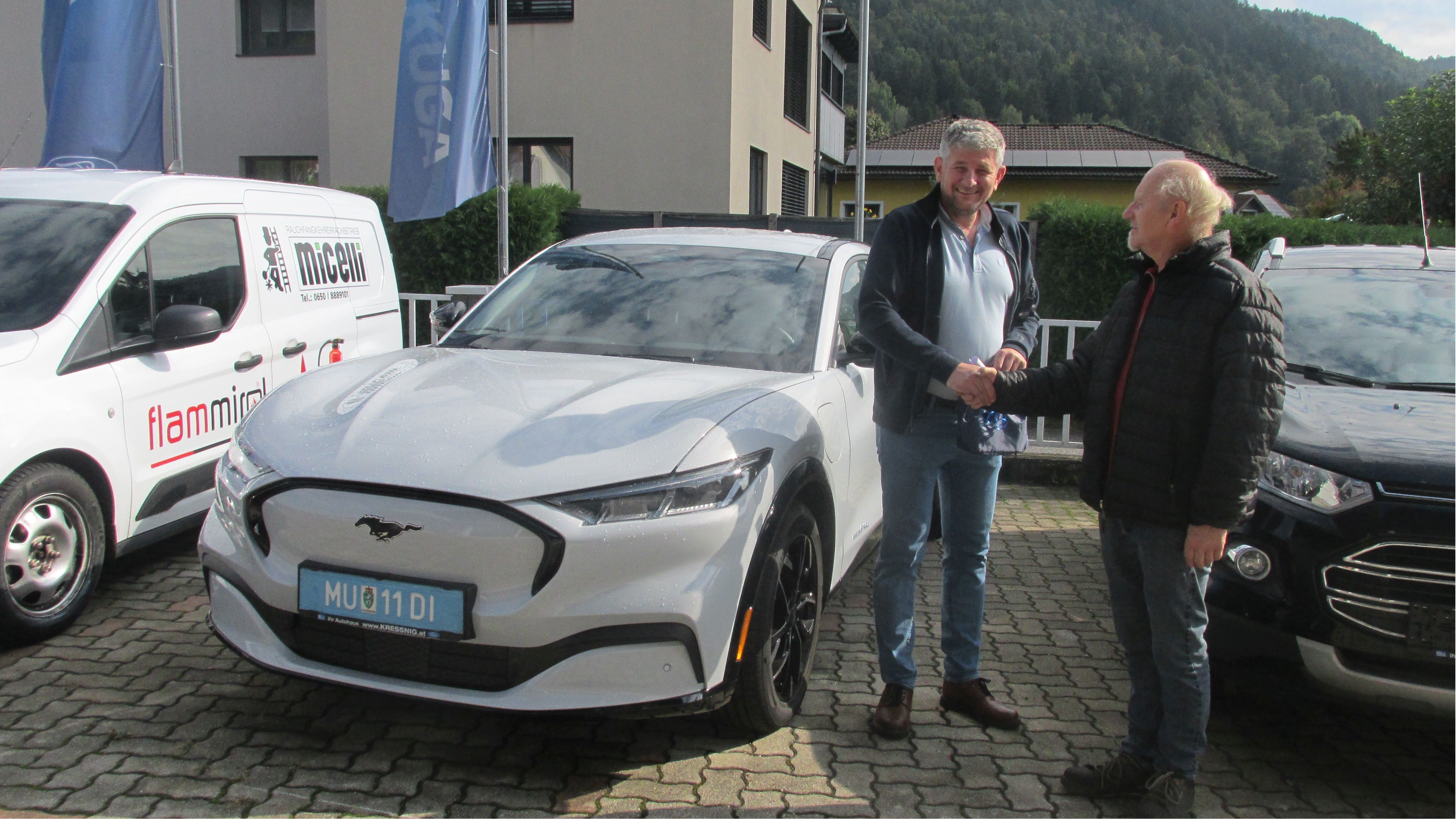 Zwei Männer schütteln sich die Hände neben einem weißen Ford Mustang Mach-E in einer urbanen Umgebung.