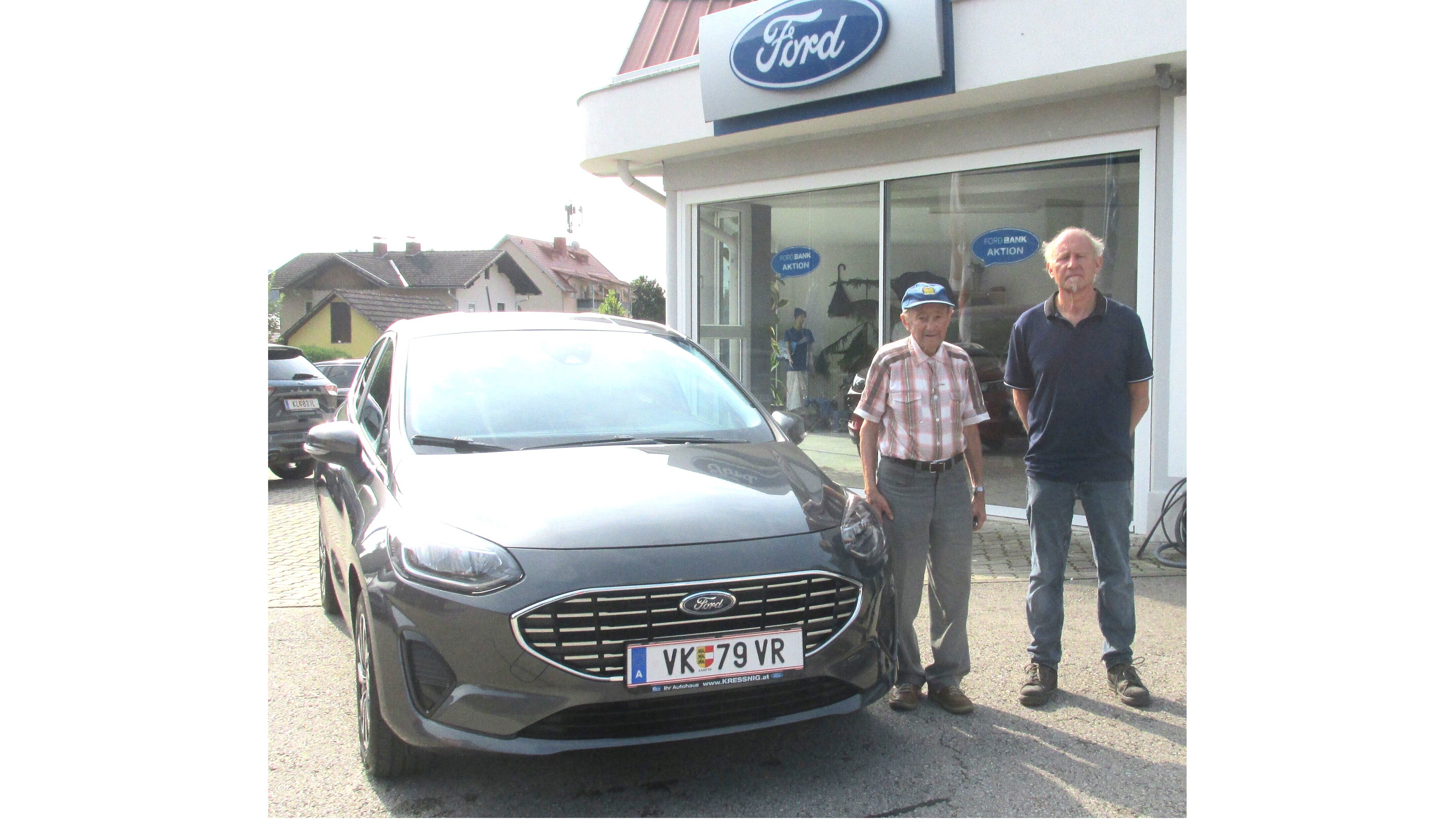 Zwei Männer stehen neben einem neuen Ford vor einem Autohaus mit Ford-Schild.