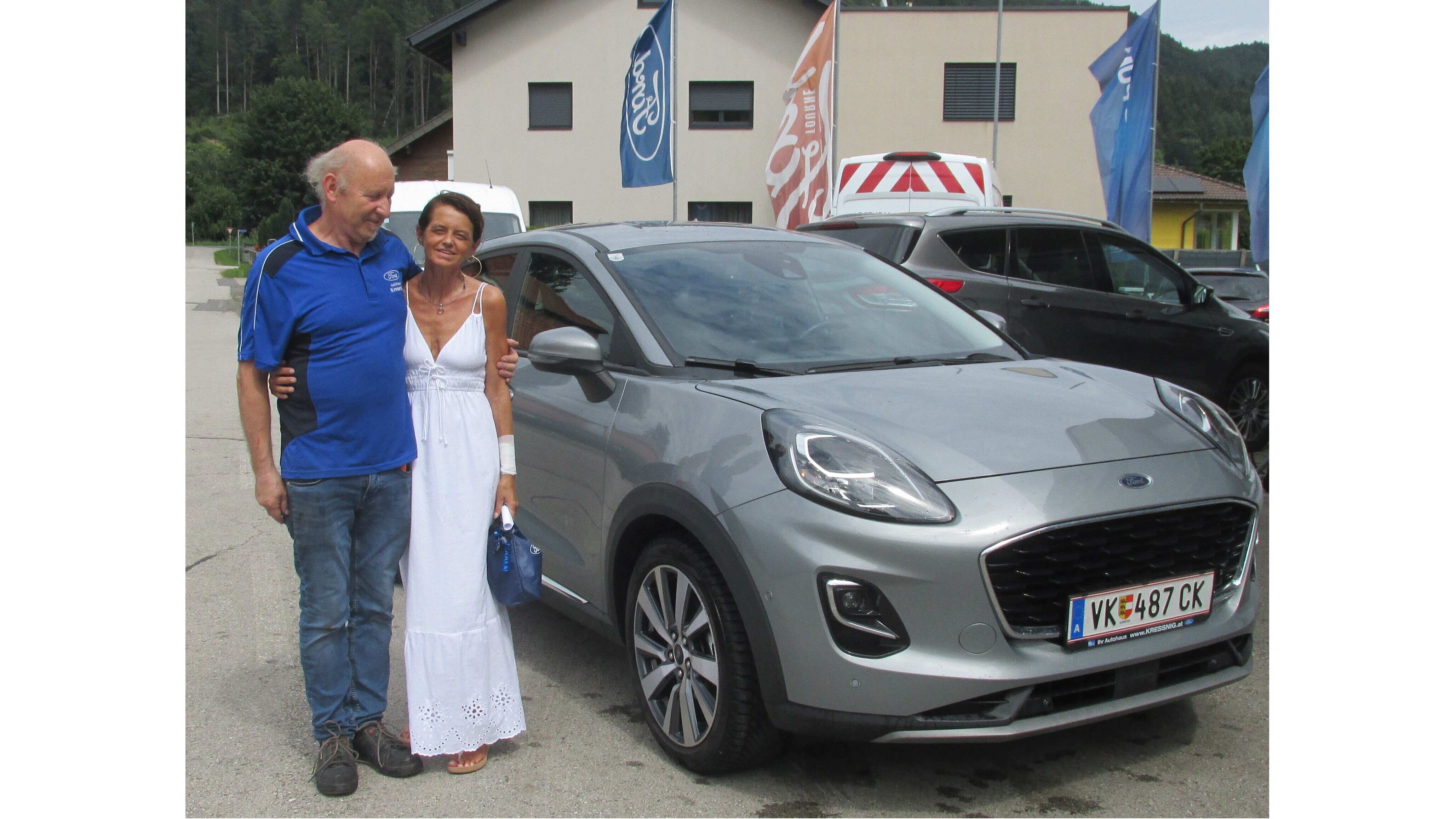 Paar posiert fröhlich neben einem silbernen Ford Puma vor einem Autohaus in der Natur.