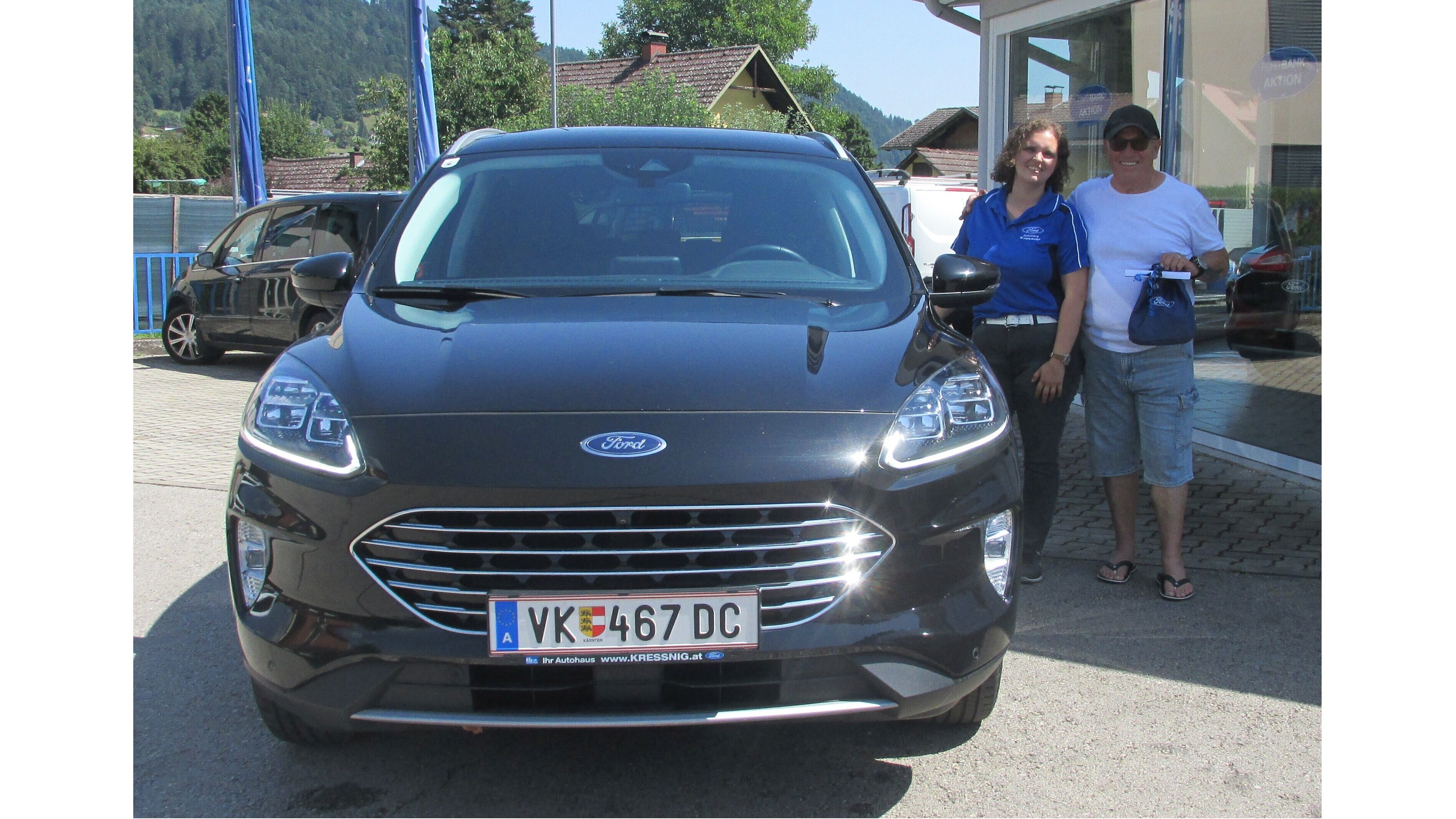 Eine Frau und ein Mann stehen neben einem schwarzen Ford SUV vor einem Autohaus, umgeben von grüner Landschaft.