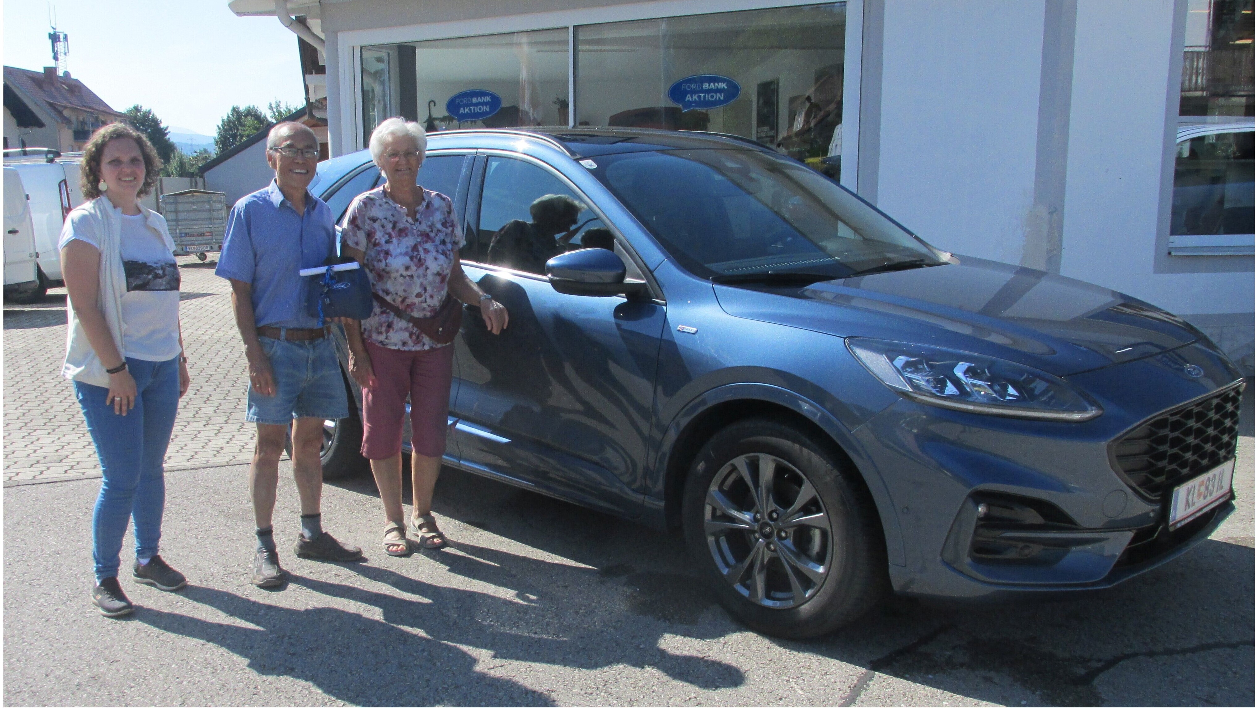 Drei Personen stehen neben einem grauen Ford Kuga vor einem Autohaus an einem sonnigen Tag.