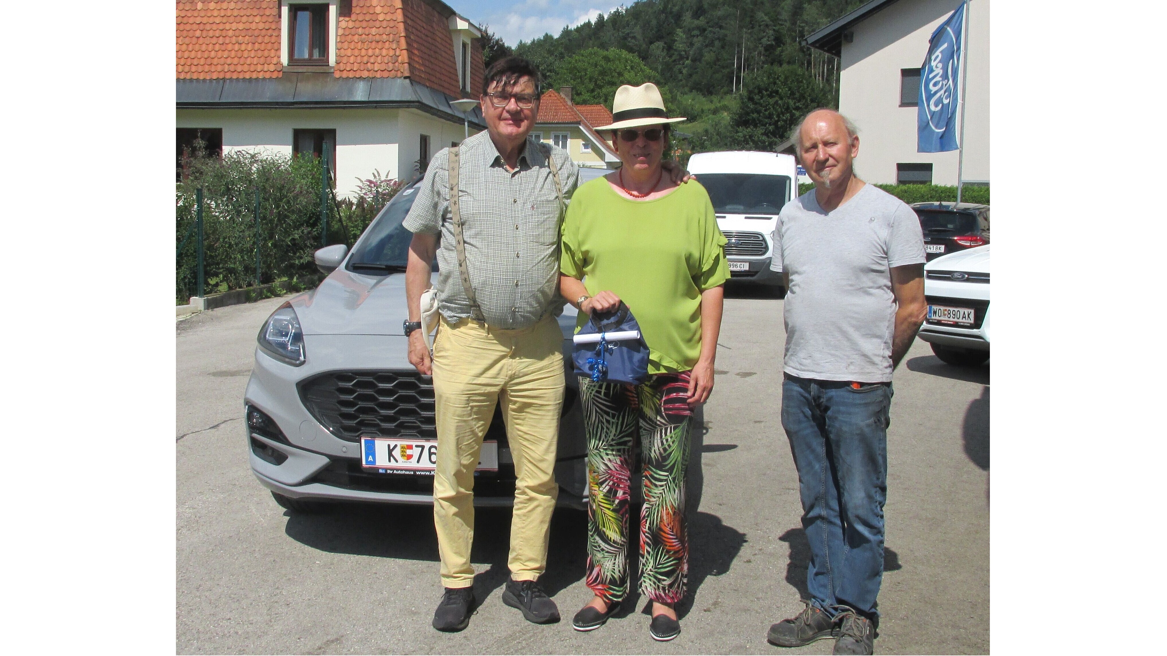 Drei Personen stehen lächelnd vor einem grauen Auto auf einer sonnigen Straße mit grüner Landschaft im Hintergrund.