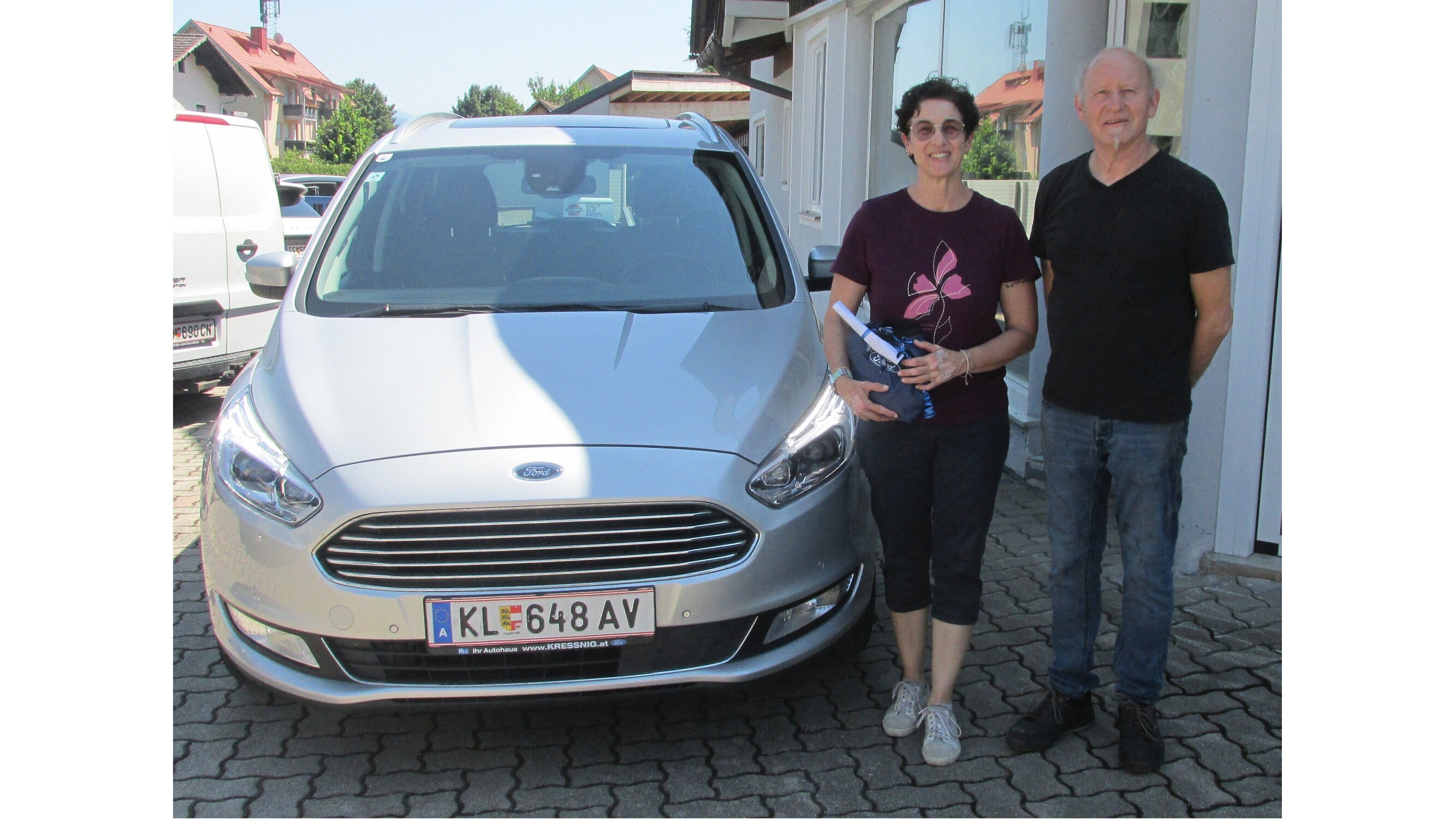 Zwei Personen stehen neben einem silbernen Ford, vor einem Gebäude. Helle Tageslichteinheit, freundliche Atmosphäre.