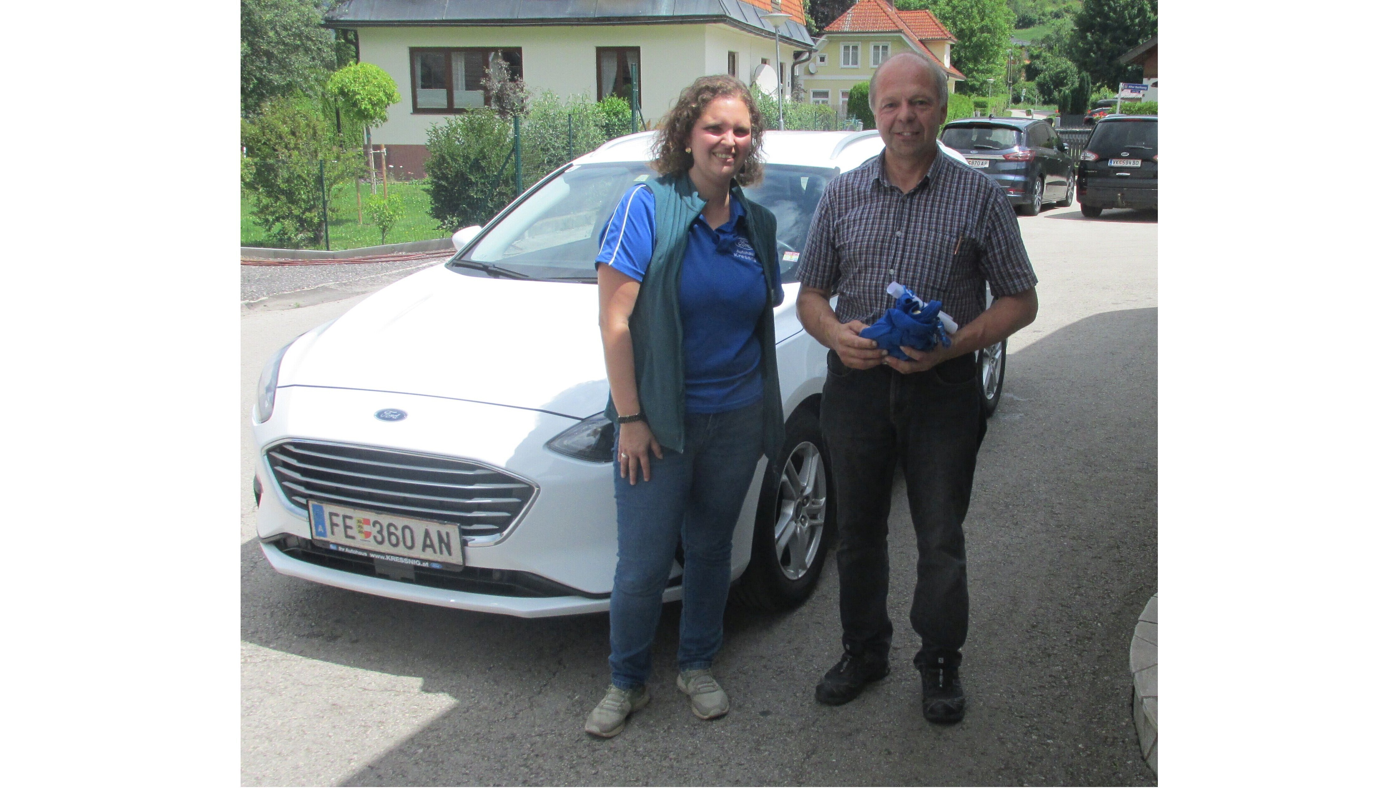 Eine Frau und ein Mann stehen vor einem weißen Ford auf einer Straße, freundlich lächelnd und miteinander sprechend.