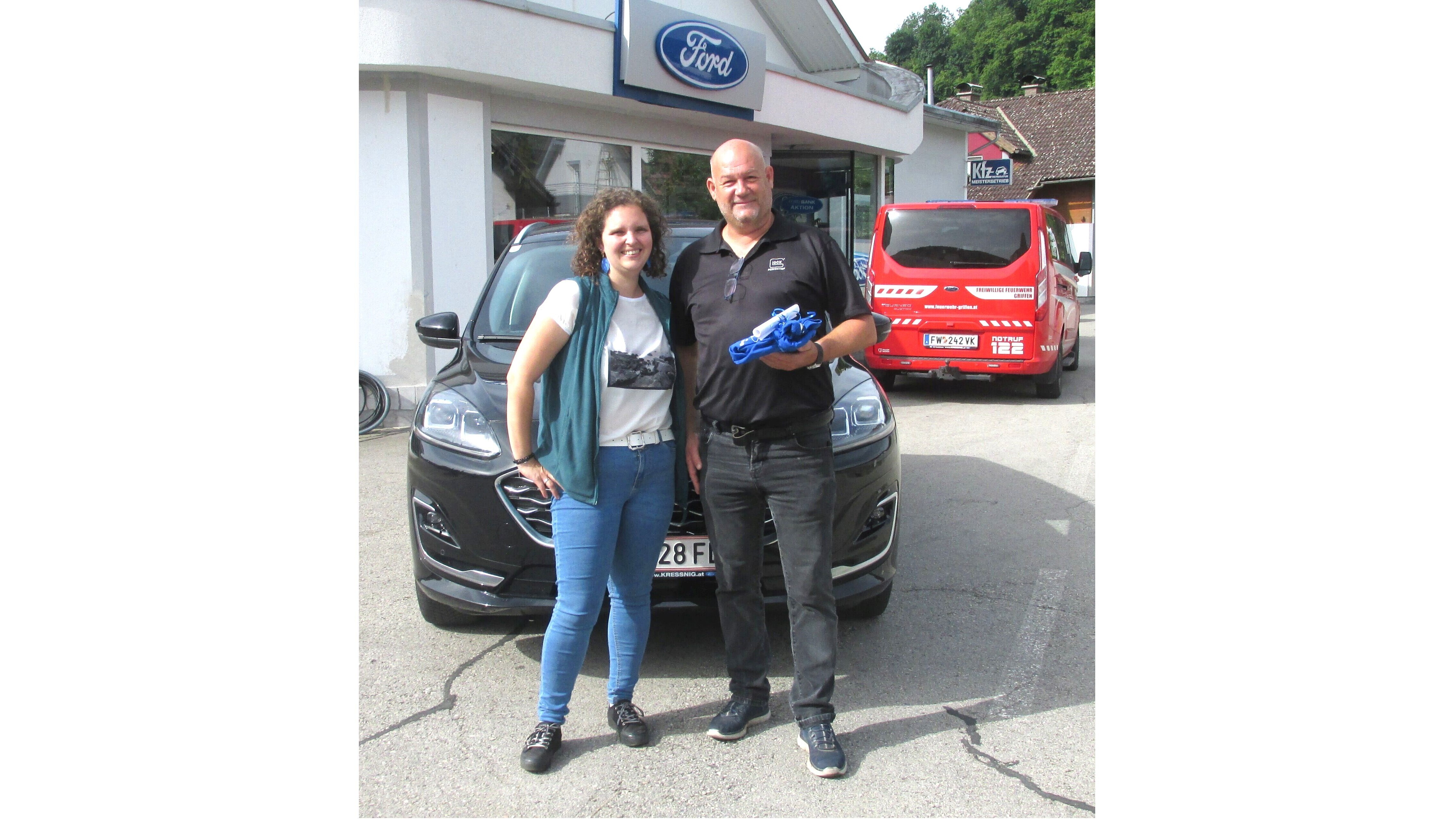 Ein Mann und eine Frau stehen vor einem schwarzen Ford-Auto an einem Autohaus. Sie lächeln, der Mann hält ein blaues Tuch.