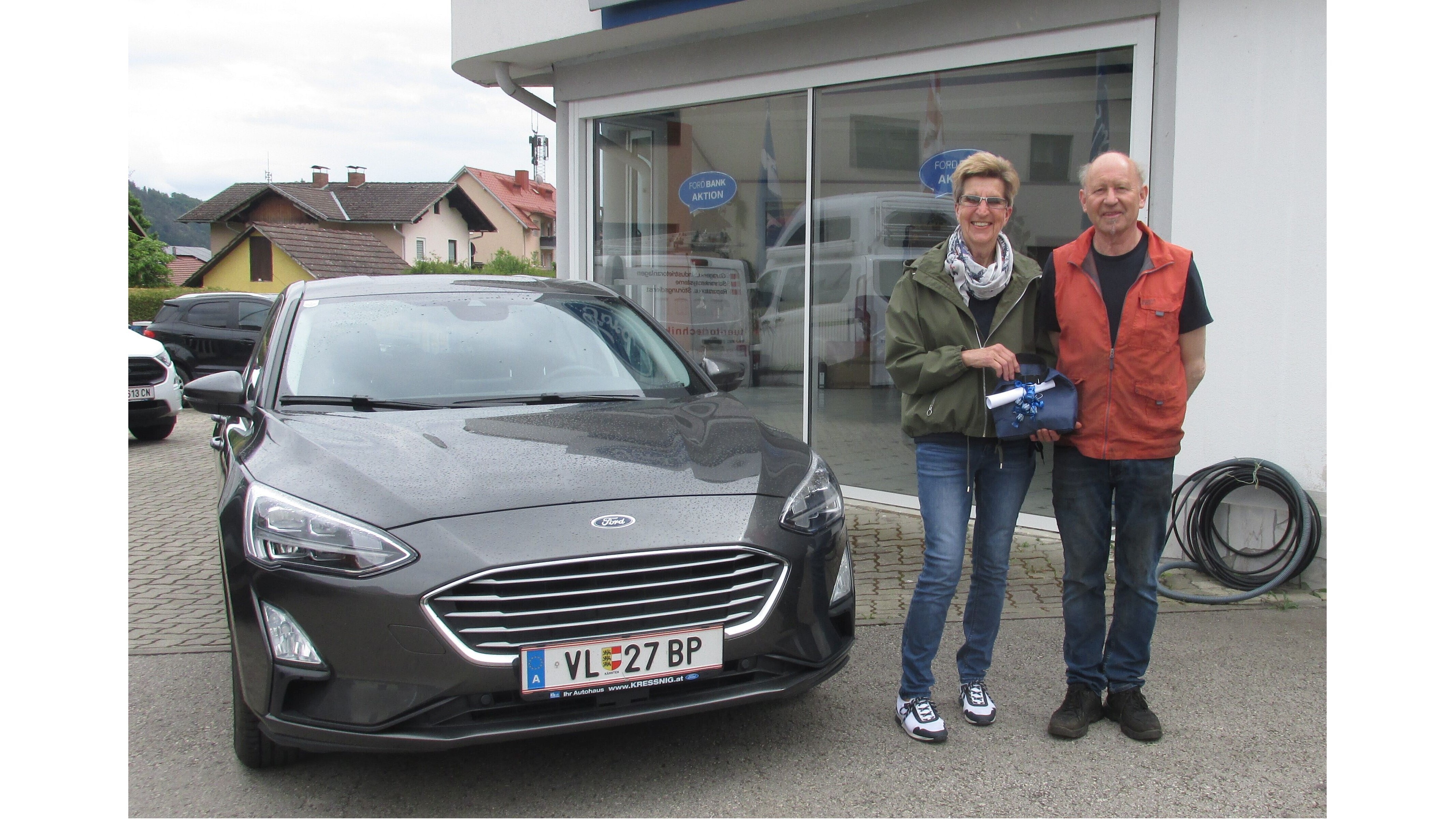 Eine Frau und ein Mann stehen neben einem grauen Auto vor einem Autohändler. Beide lächeln in die Kamera.