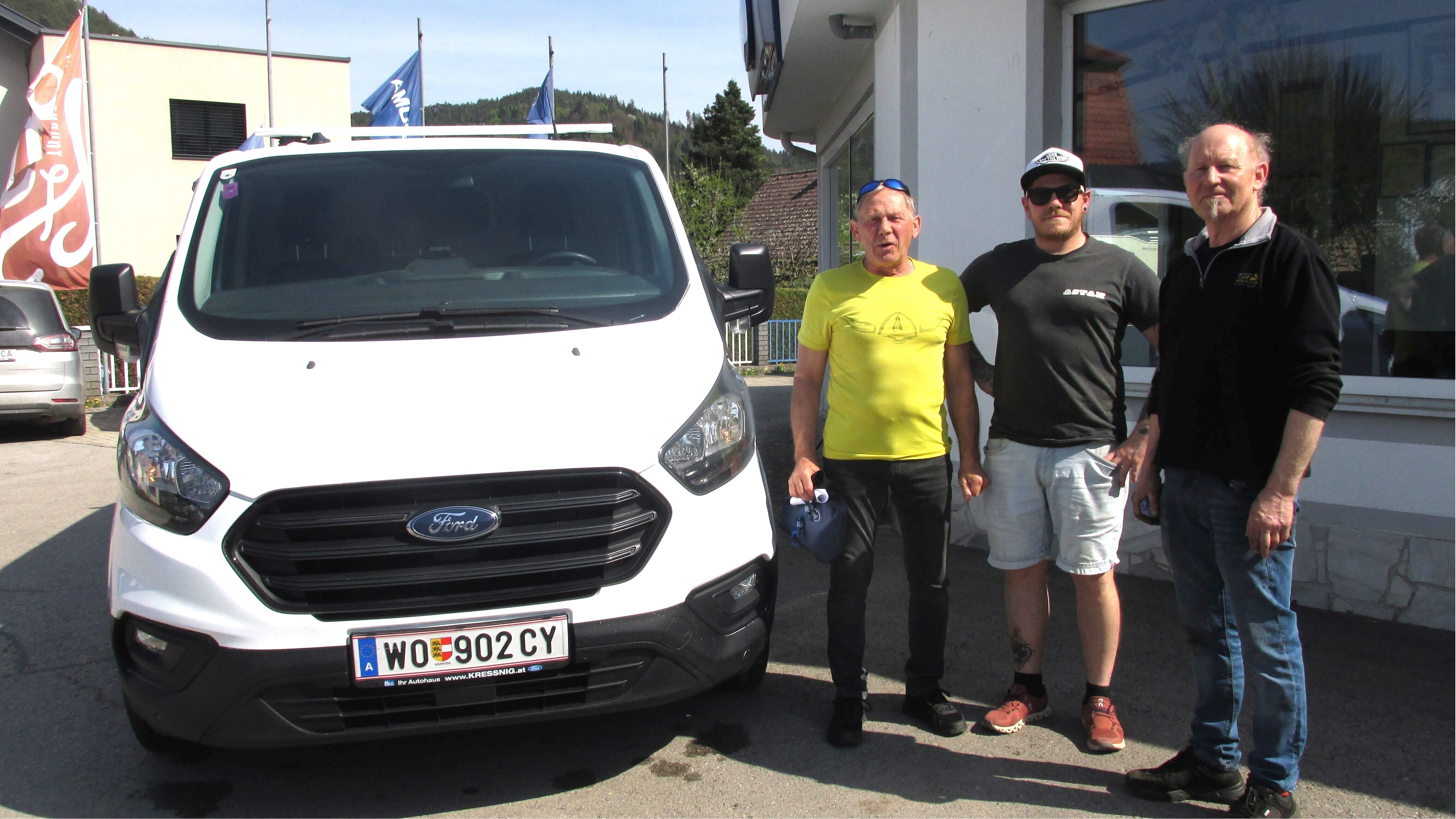 Drei Männer stehen neben einem weißen Ford Transporter vor einem Autohaus, sonniger Tag, Berge im Hintergrund.