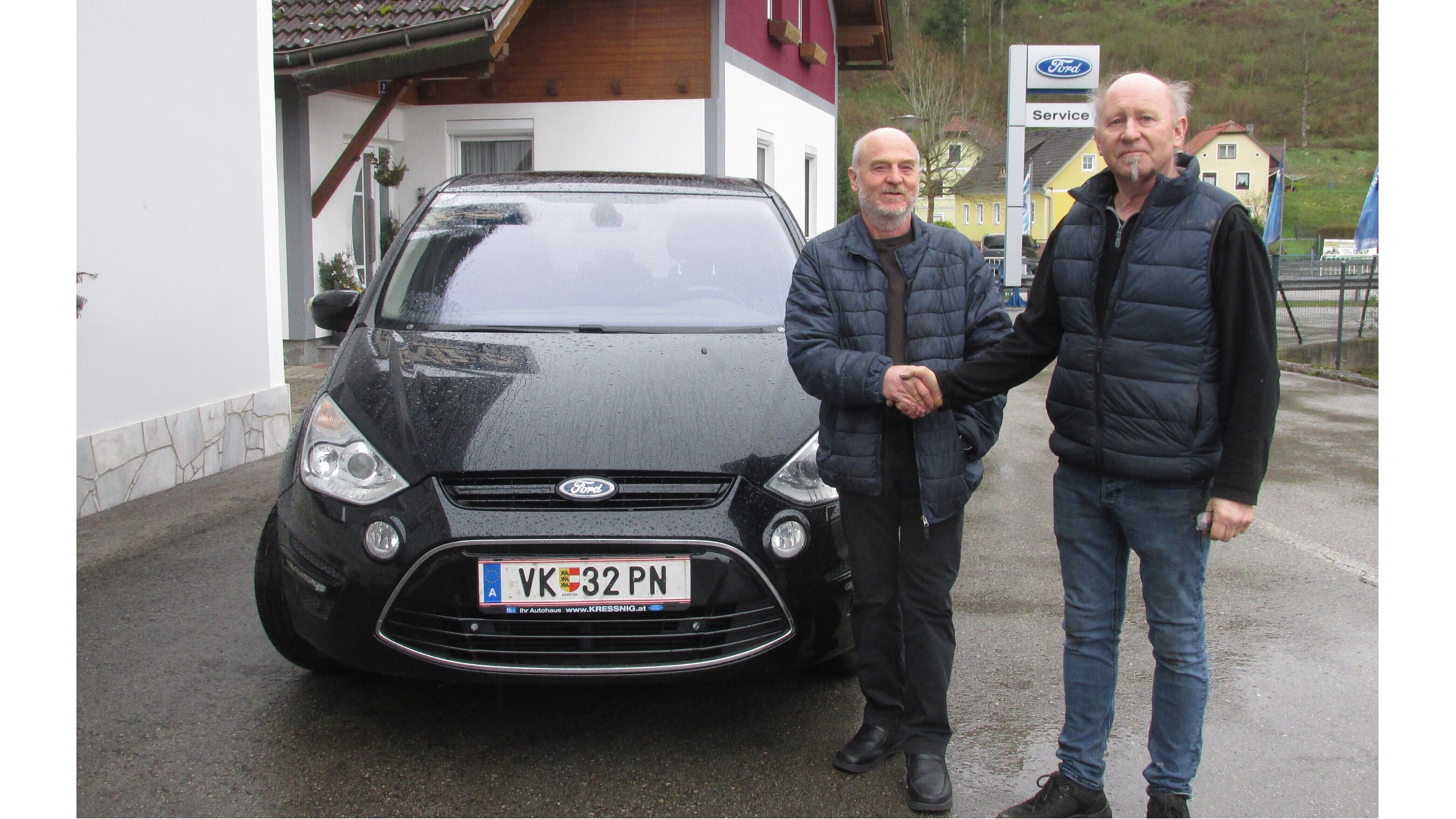 Zwei Männer schütteln sich die Hände vor einem schwarzen Ford und einem Autohaus im Hintergrund.