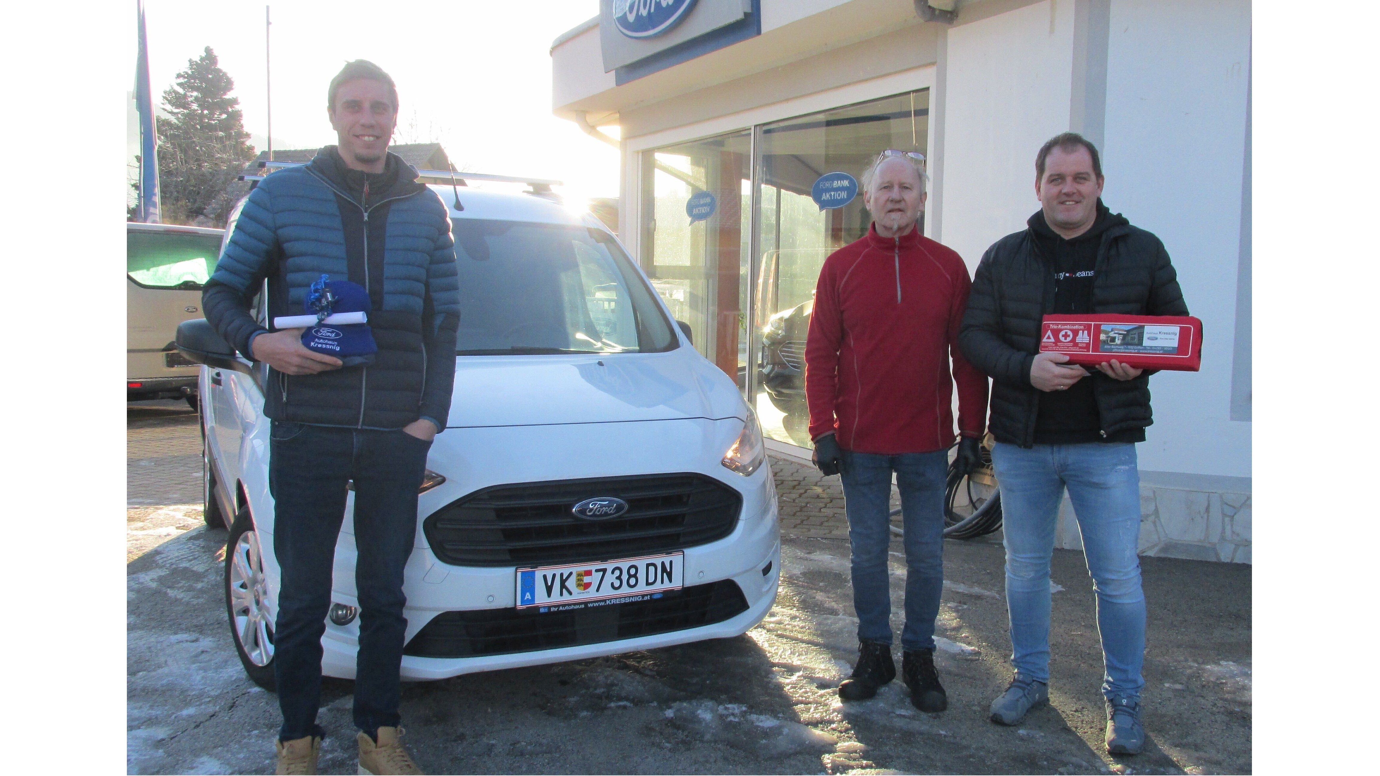 Drei Männer stehen neben einem weißen Ford im Freien, sie halten Geschenke und posieren vor einem Ford-Händler.