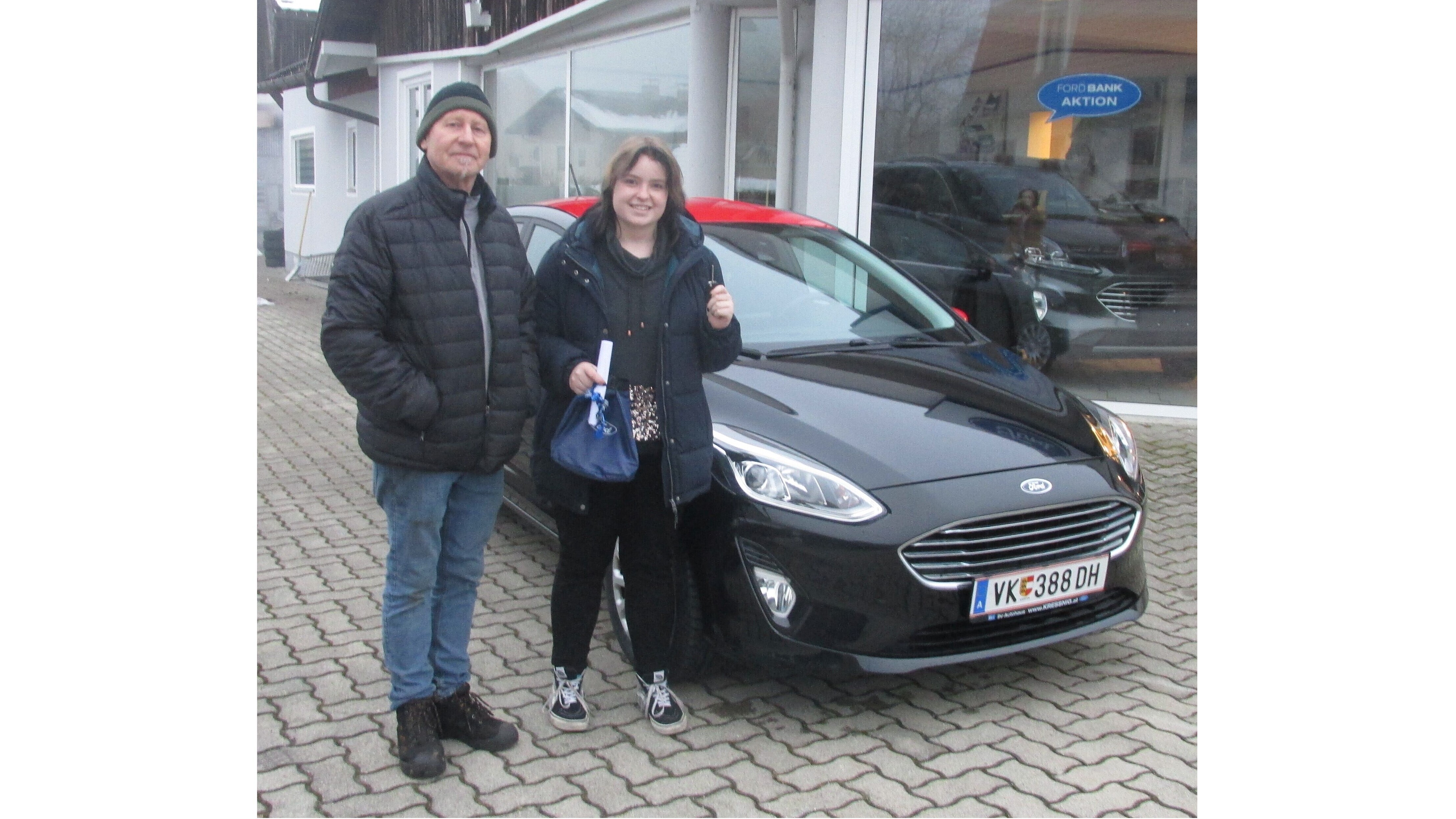Ein Mann und eine Frau posieren lächelnd neben einem schwarzen Ford. Sie stehen vor einem Autohaus.
