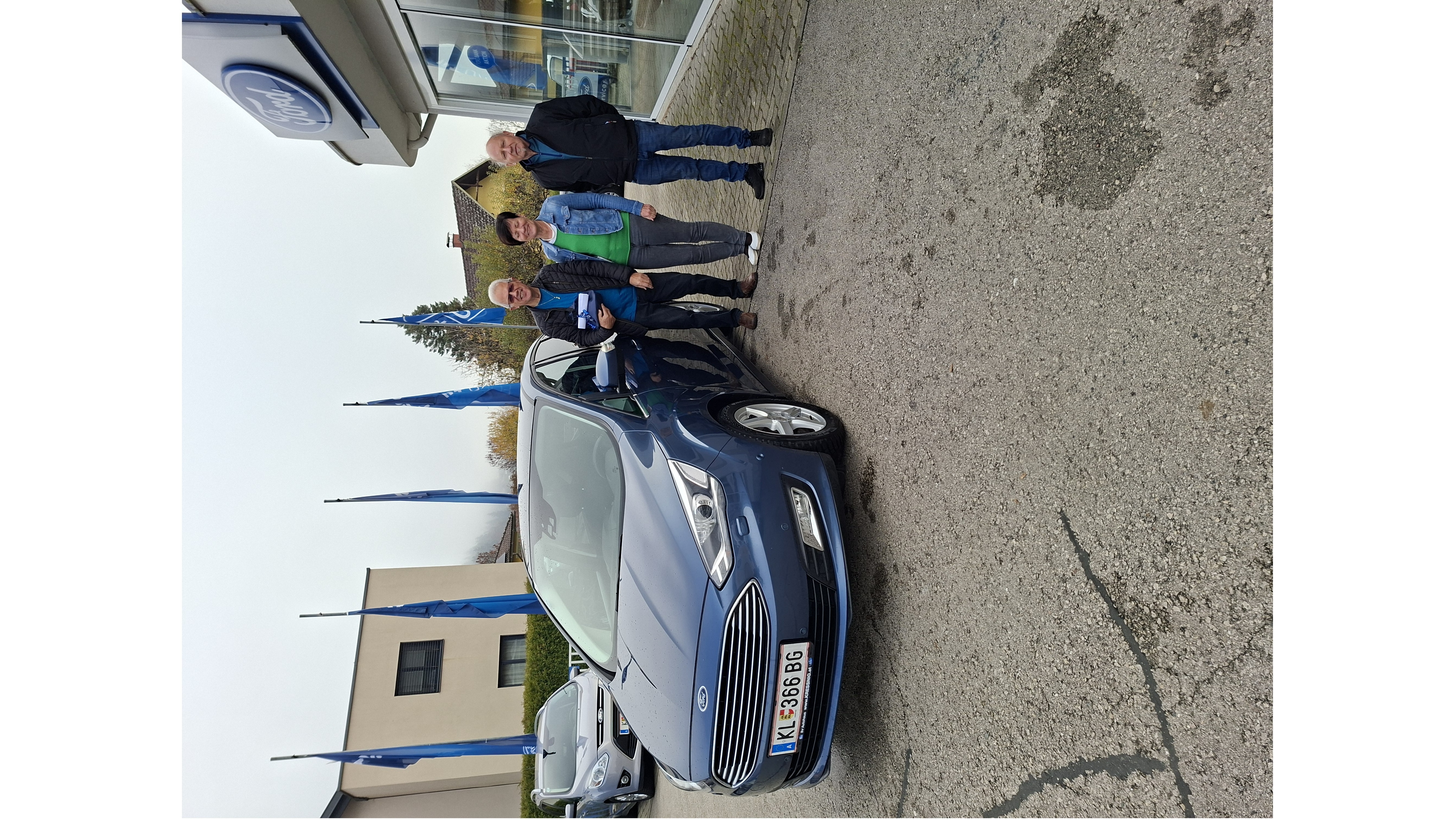 Drei Personen stehen vor einem blauen Ford, umgeben von einem Autohaus mit Flaggen.