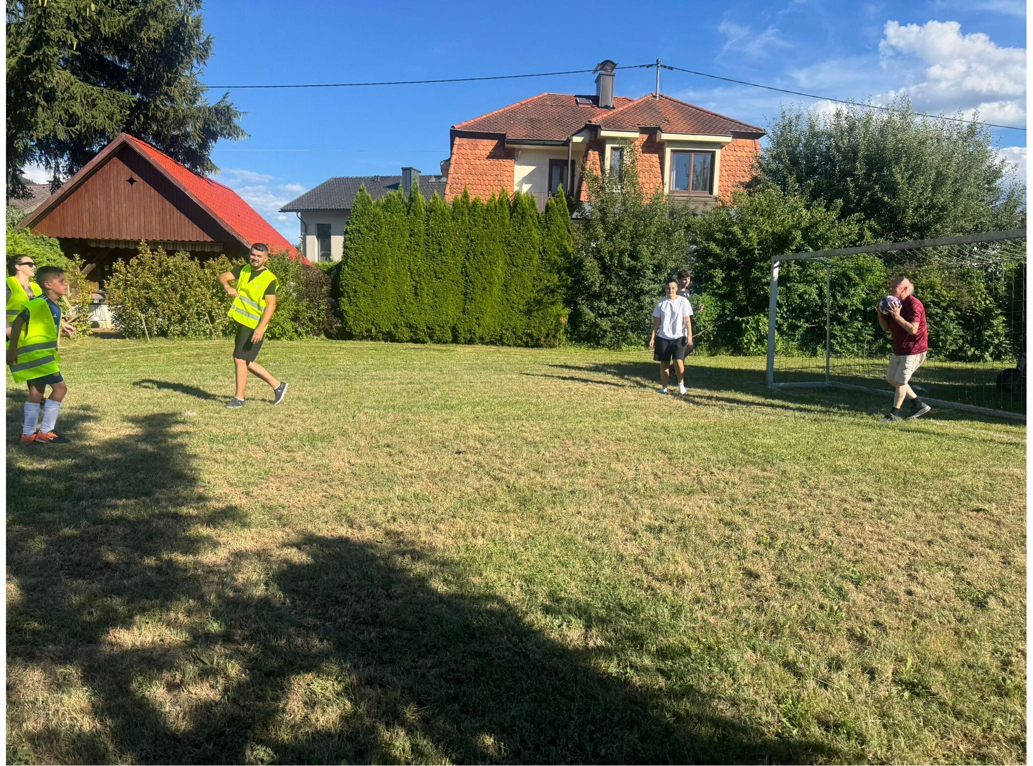 Jugendliche in Warnwesten spielen Fußball auf einer Wiese vor einem Haus mit einem Tor im Hintergrund.