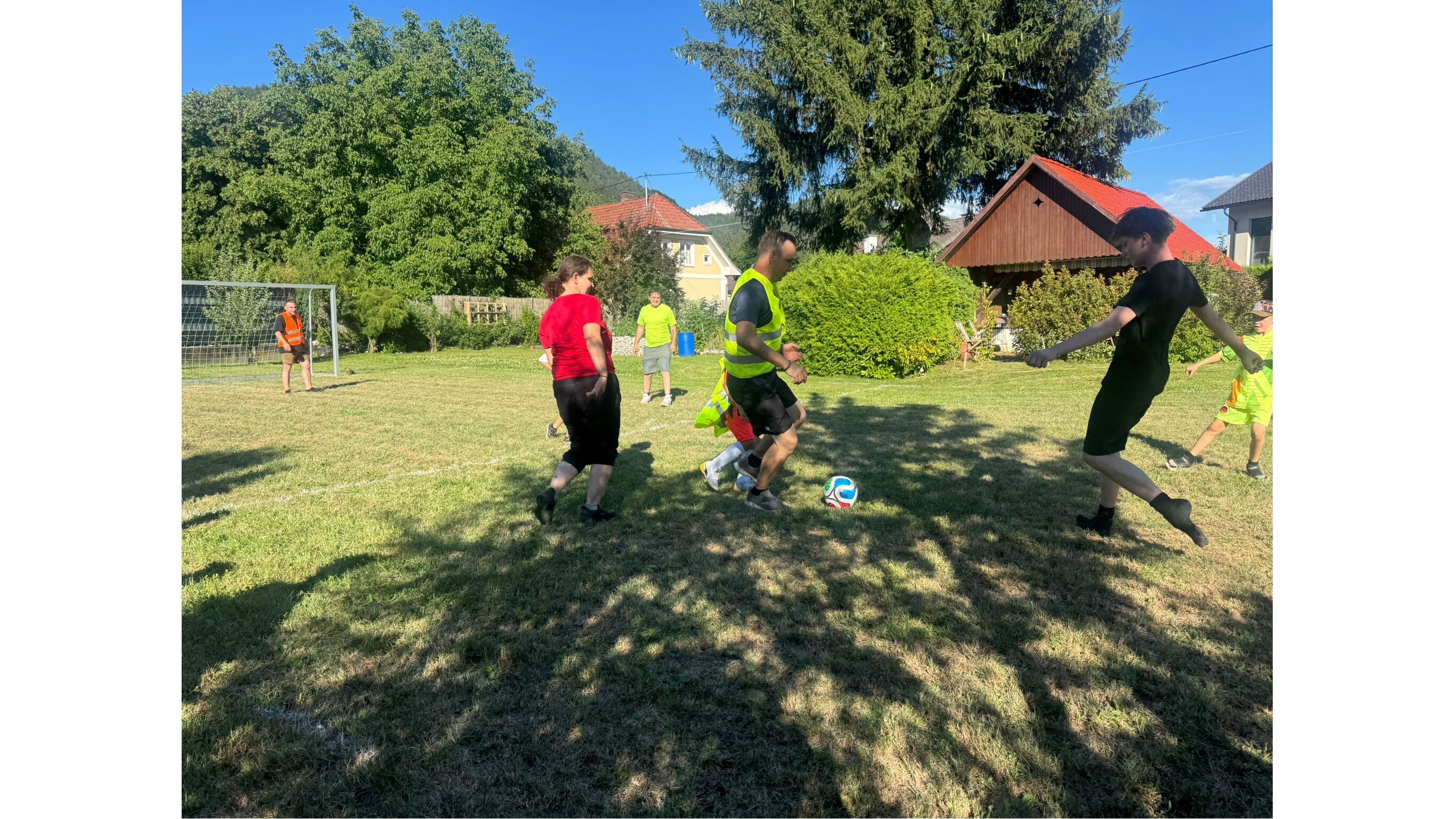 Kinder und Erwachsene spielen Fußball auf einem grünen Rasenfeld, umgeben von Bäumen und Häusern.