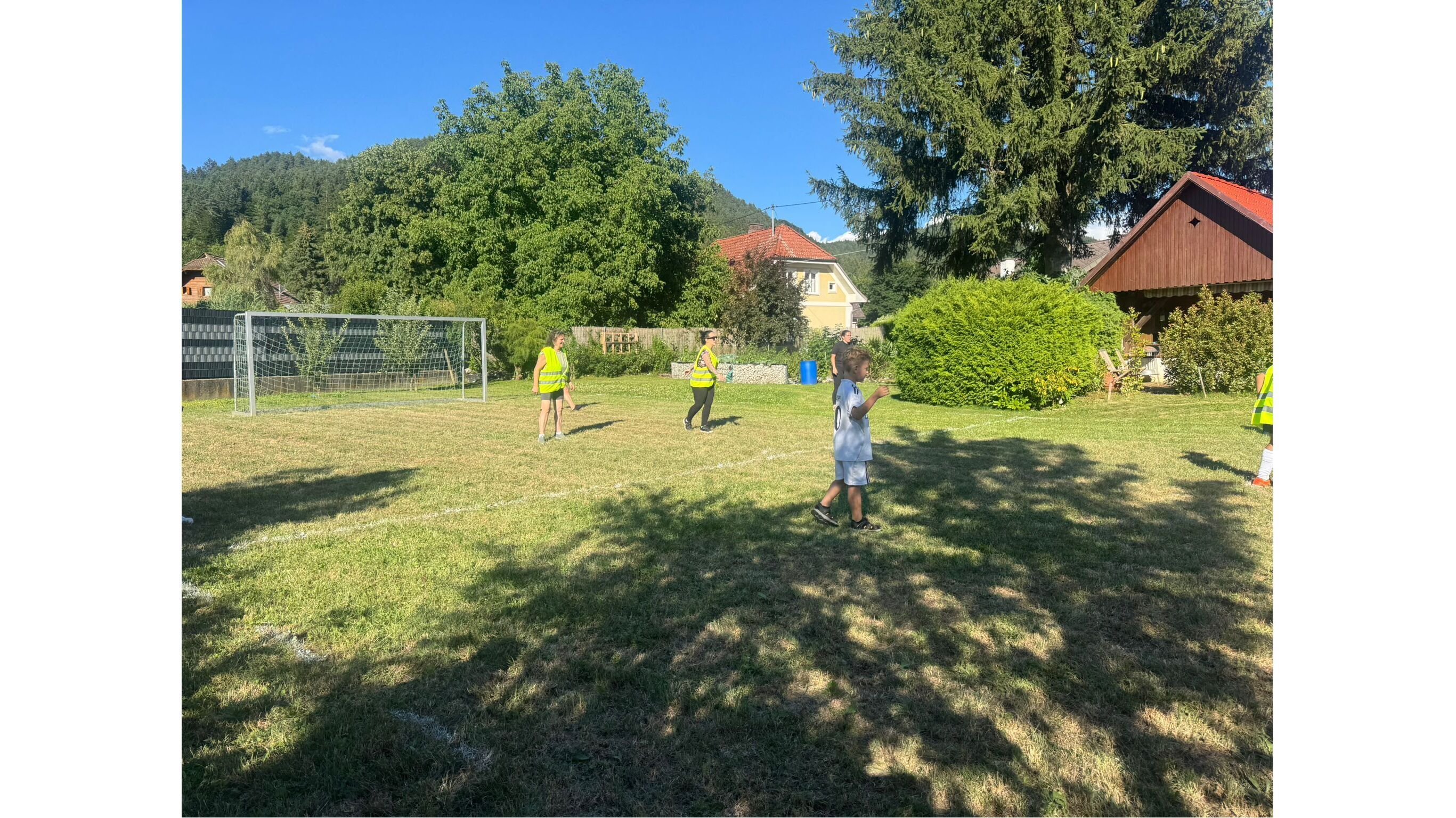 Kinder spielen Fußball auf einem Rasenplatz mit Tor und Bäumen im Hintergrund, bei schönem Wetter.