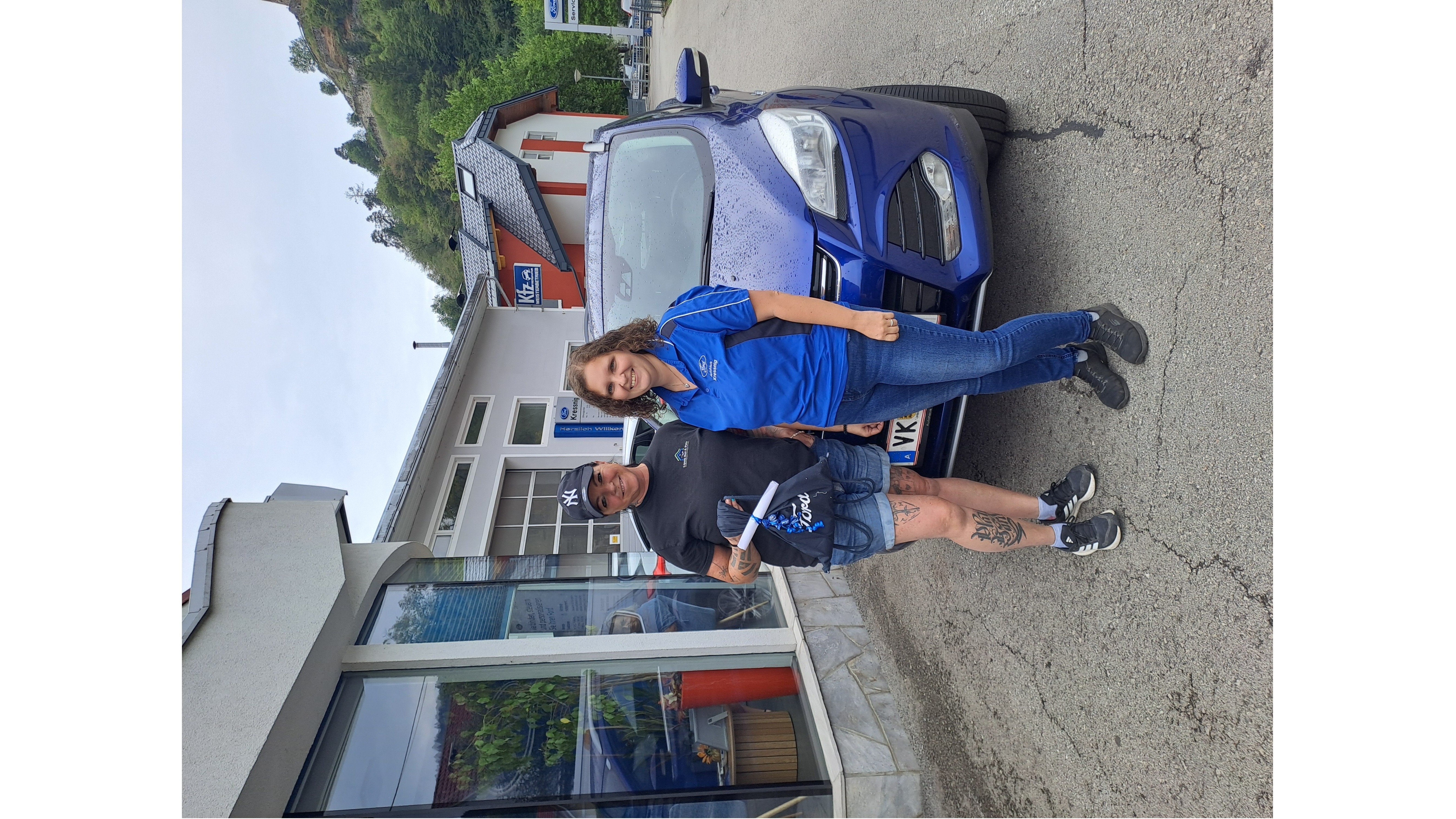 Zwei Personen stehen lächelnd vor einem blauen Auto in einer Werkstattansicht, umgeben von Gebäuden.