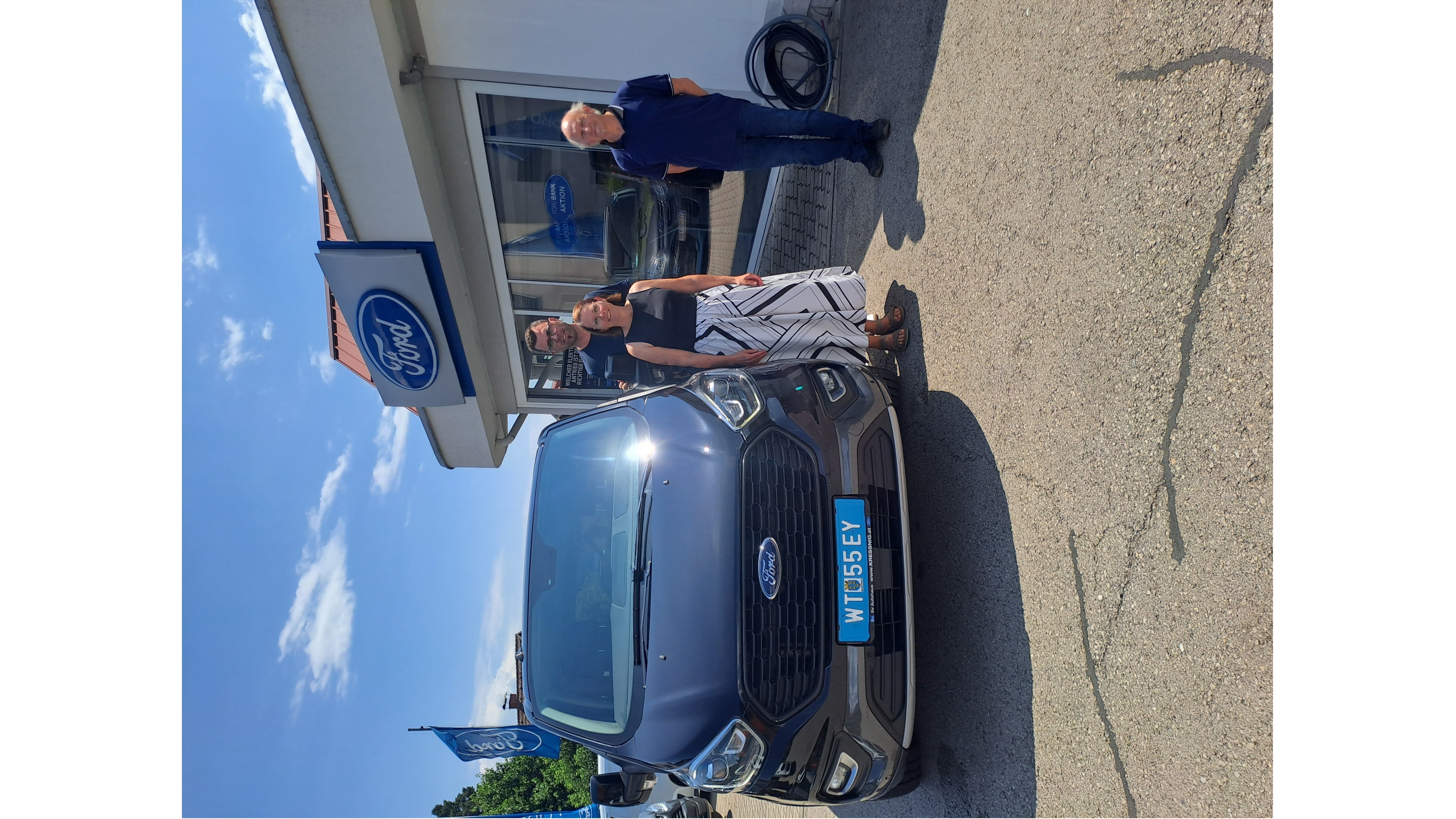 Ein Mann und eine Frau stehen vor einem Ford Fahrzeug vor einem Ford Autohaus unter blauem Himmel.