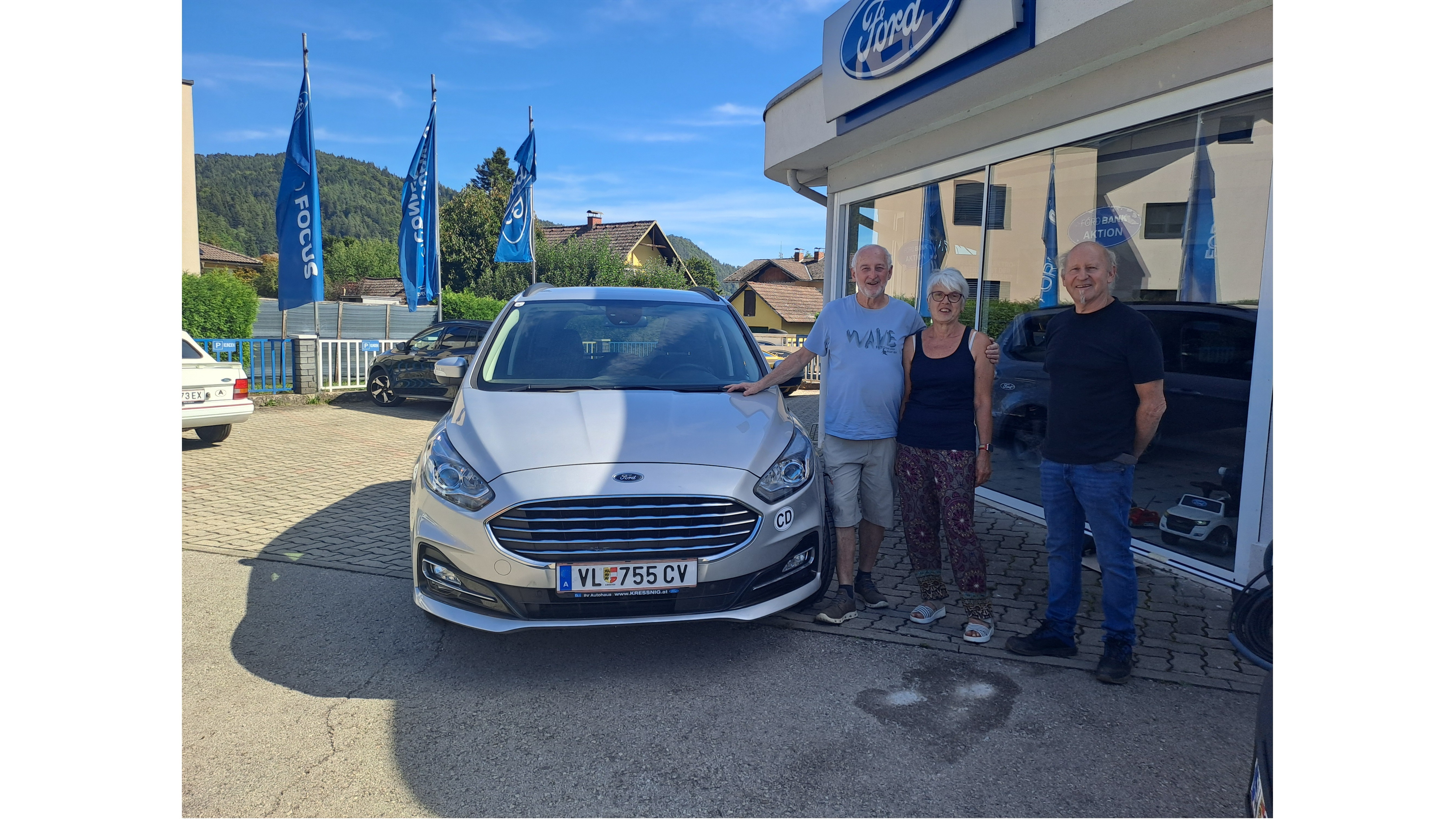 Drei lächelnde Personen stehen neben einem neuen Ford-Wagen vor einem Autohaus mit Fahnen im Hintergrund.