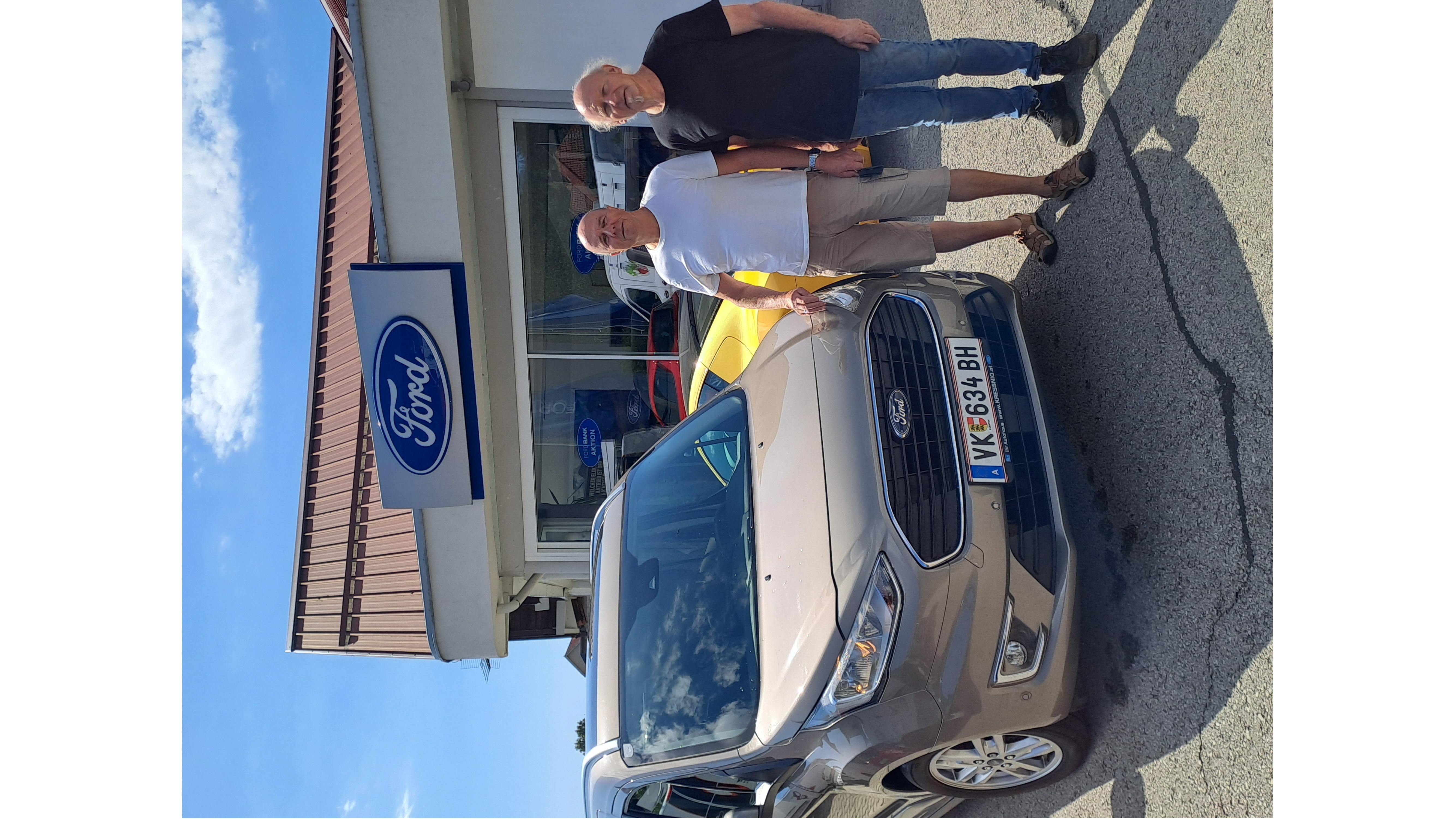 Zwei Männer stehen neben einem Ford-Fahrzeug vor einem Ford-Händler in einer sonnigen Umgebung.