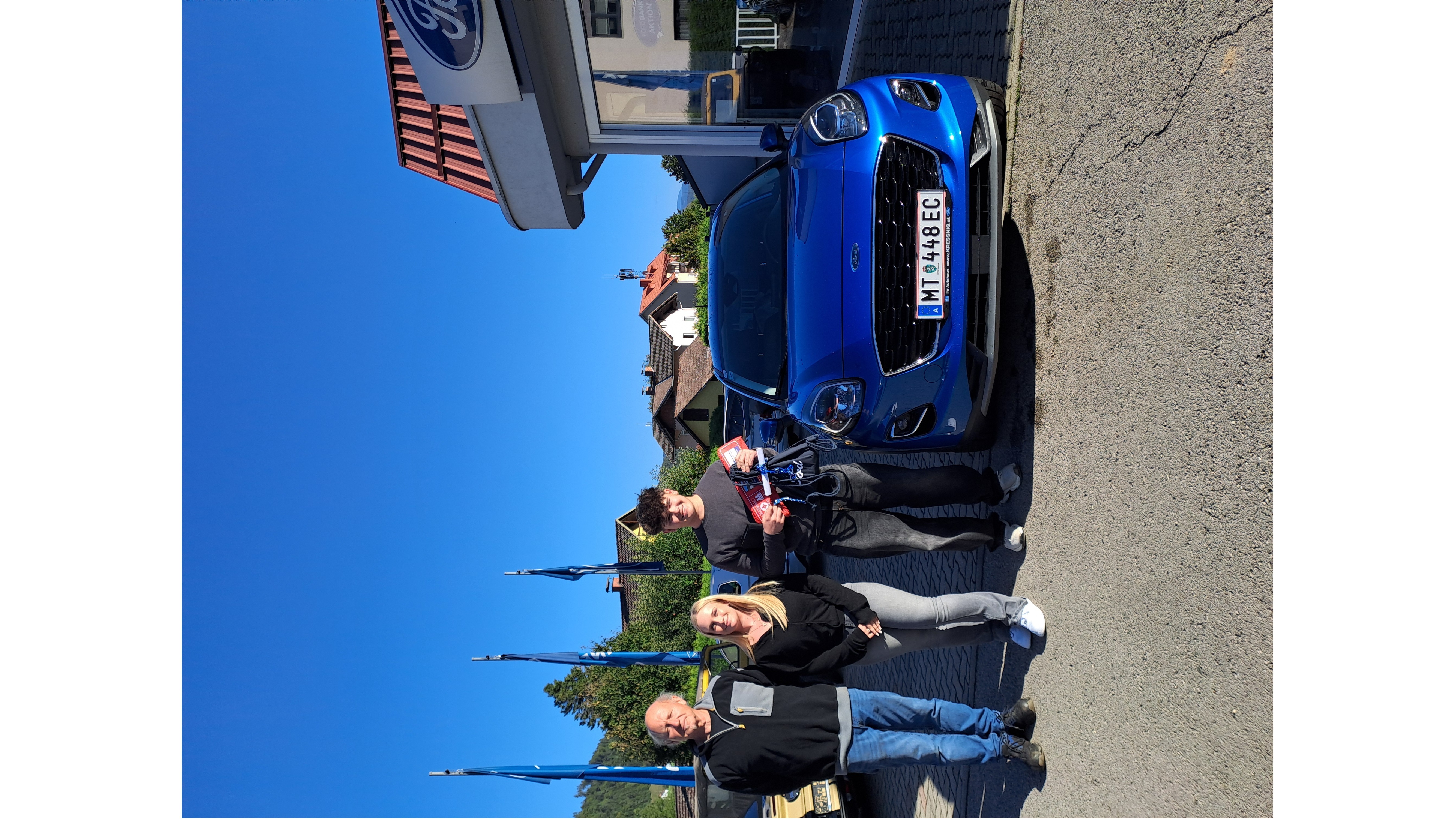Drei Personen posieren mit einem blauen Ford-Auto vor einem Autohaus bei klarem Himmel.