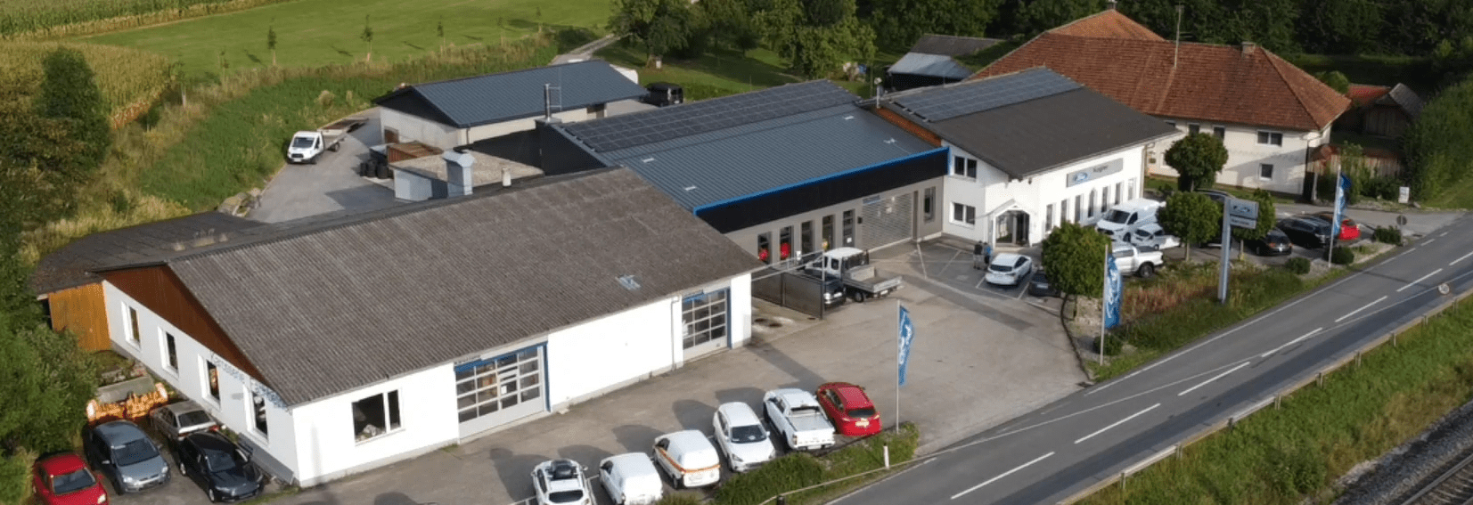 Luftaufnahme eines Firmengebäudes mit mehreren Autos und einer Umgebung aus Grünflächen und Wohnhäusern.