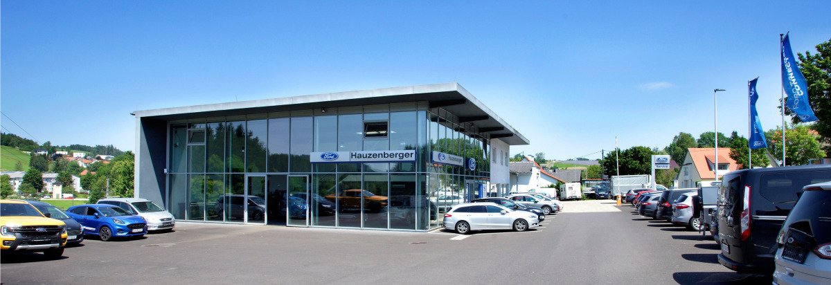 Autohaus Hauzenberger mit moderner Glasfassade und Parkplätzen, umgeben von grüner Landschaft.