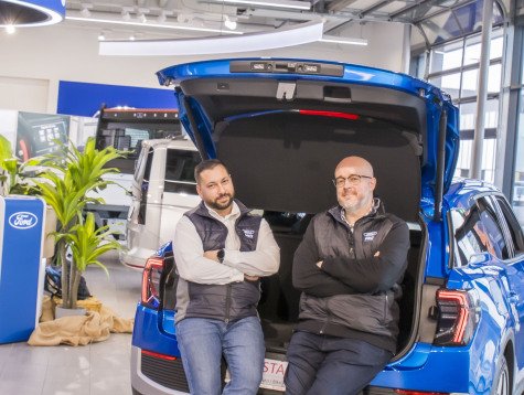 Zwei Männer sitzen lässig auf dem Heck eines blauen Ford-Fahrzeugs in einem modernen Autohaus.