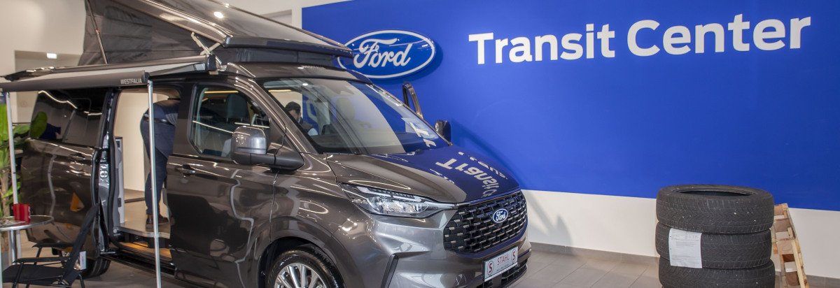 Ein grauer Ford Transit im Ford Transit Center mit offenem Dachzelt und einer blauen Wand im Hintergrund.