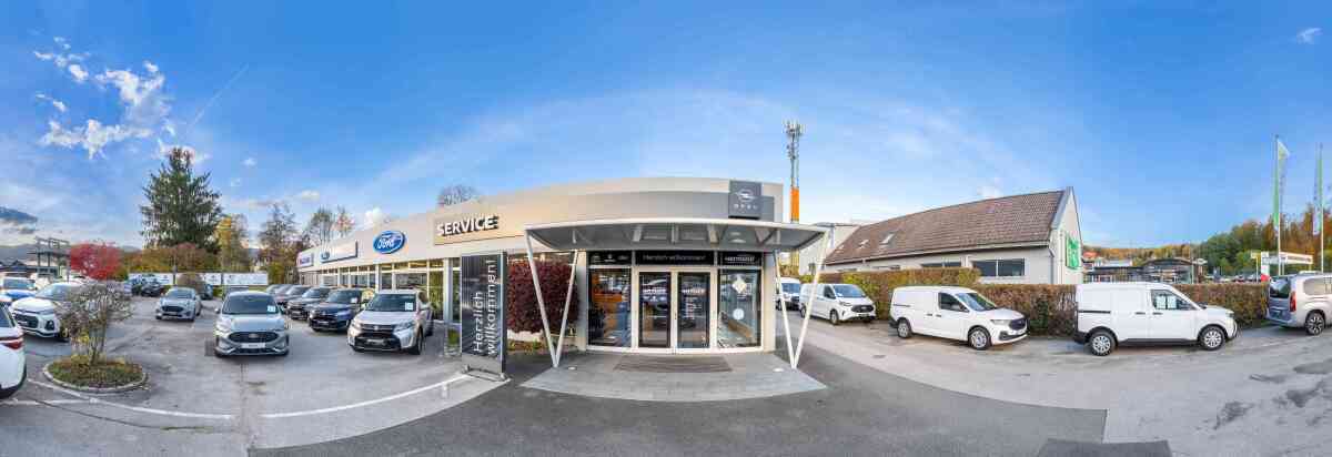 Panoramaaufnahme eines Autohauses mit Parkplätzen und Autos unter blauem Himmel.