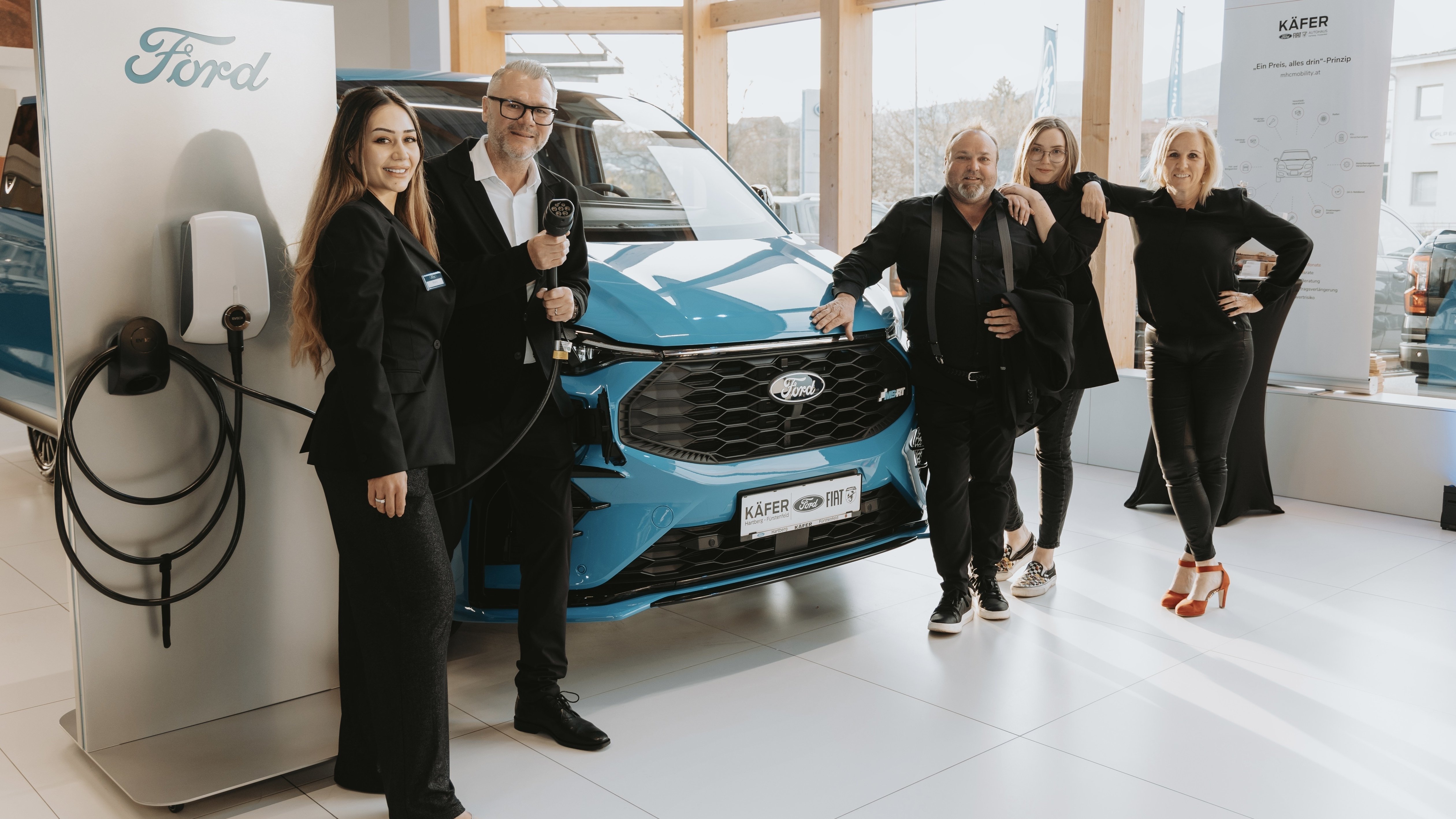 Fünf Personen posieren vor einem blauen Ford Auto und einer Ladestation in einem modernen Ausstellungsraum.