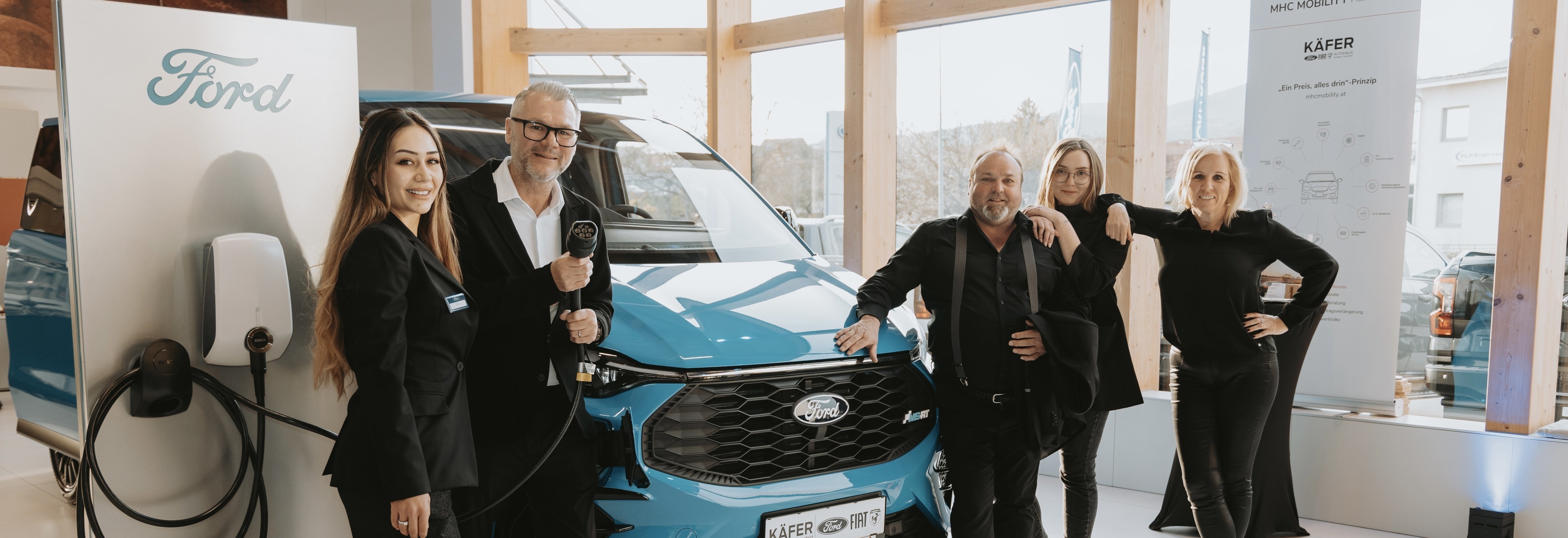 Gruppenszenen mit fünf Personen vor einem blauen Ford-Fahrzeug und einem Ladegerät, in einem modernen Ausstellungsraum.