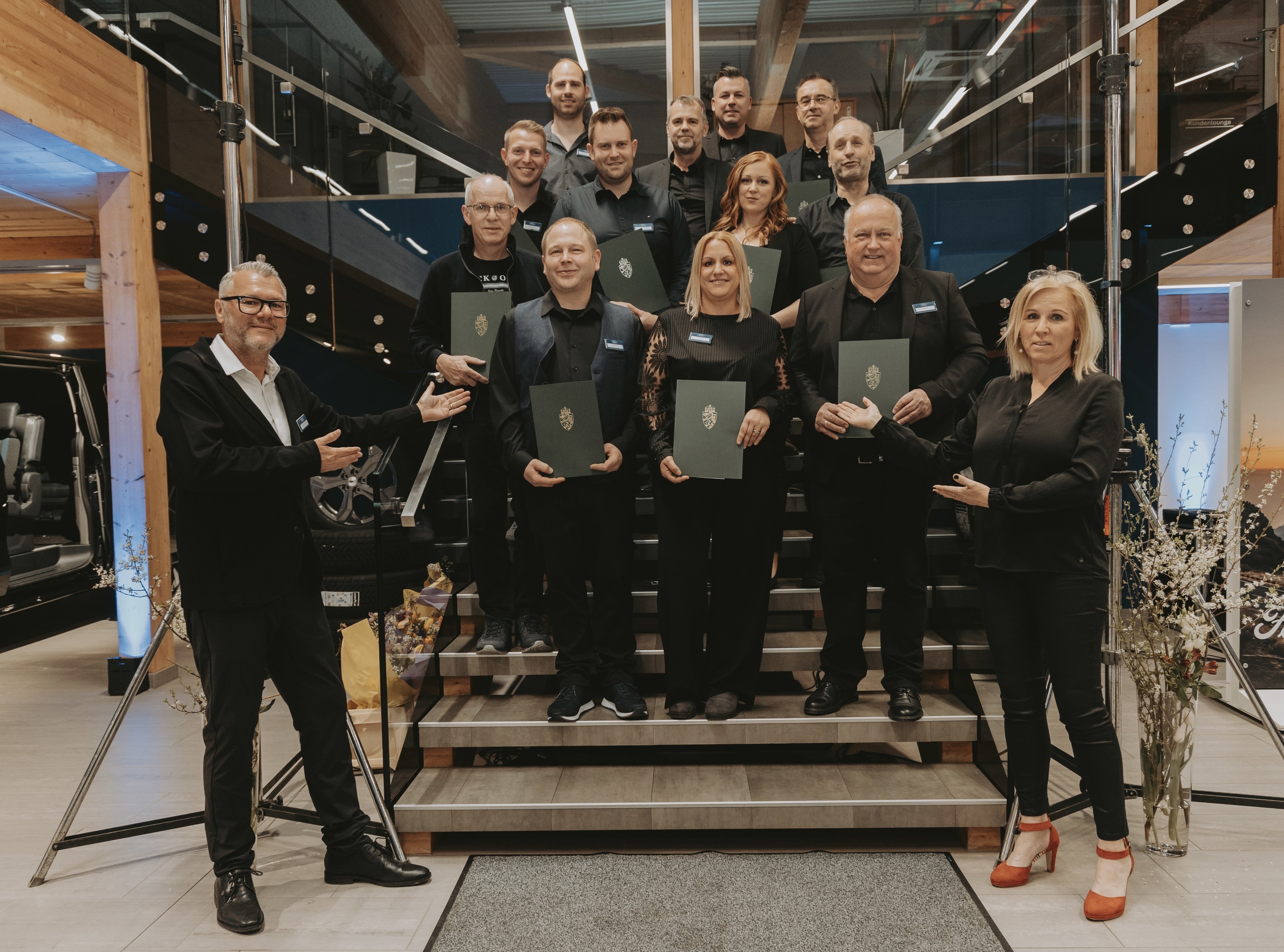 Gruppenfoto von Mitarbeitern in Schwarz, posierend mit Urkunden auf einer Treppe in einem modernen Raum.