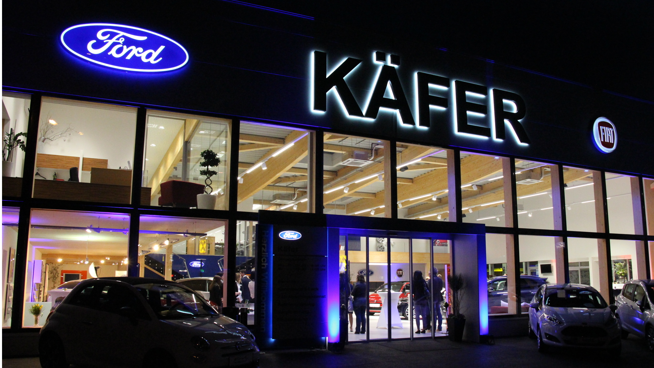 Ford Autohaus Käfer bei Nacht mit beleuchtetem Logo und Autos in der Ausstellung.