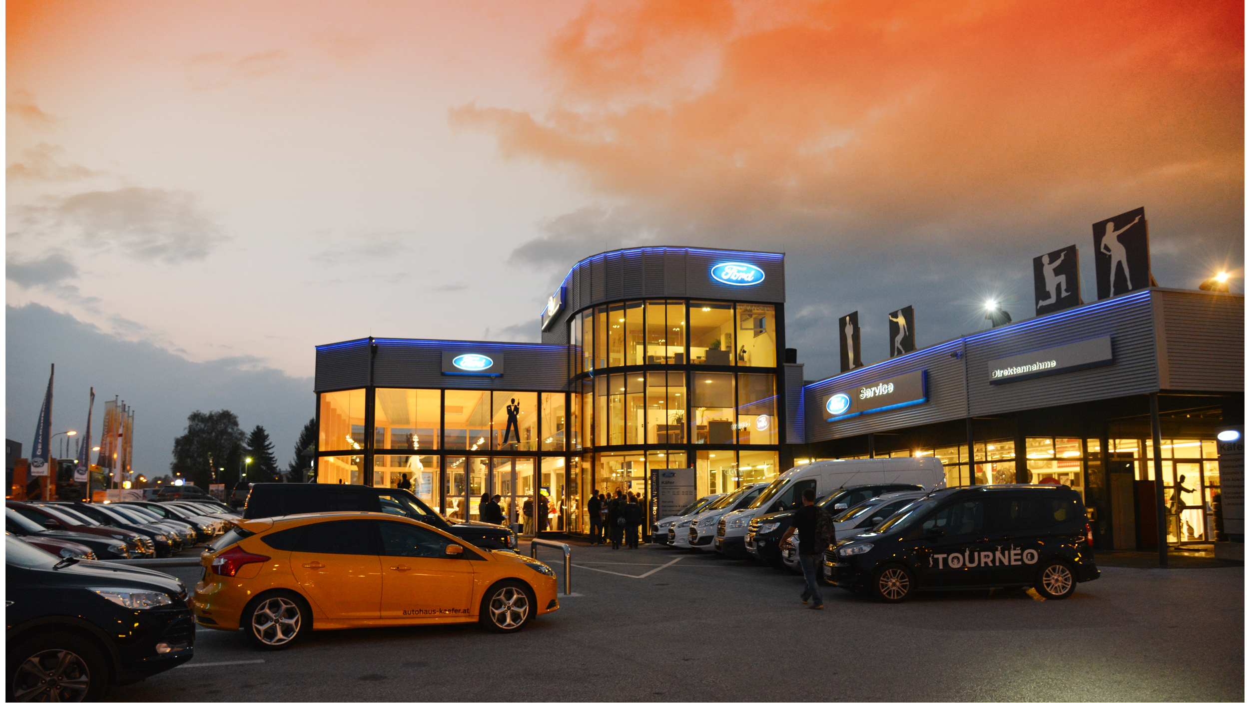 Moderne Ford-Filiale bei Sonnenuntergang, zahlreiche Autos vor dem Gebäude, beleuchtete Werbeanzeigen.