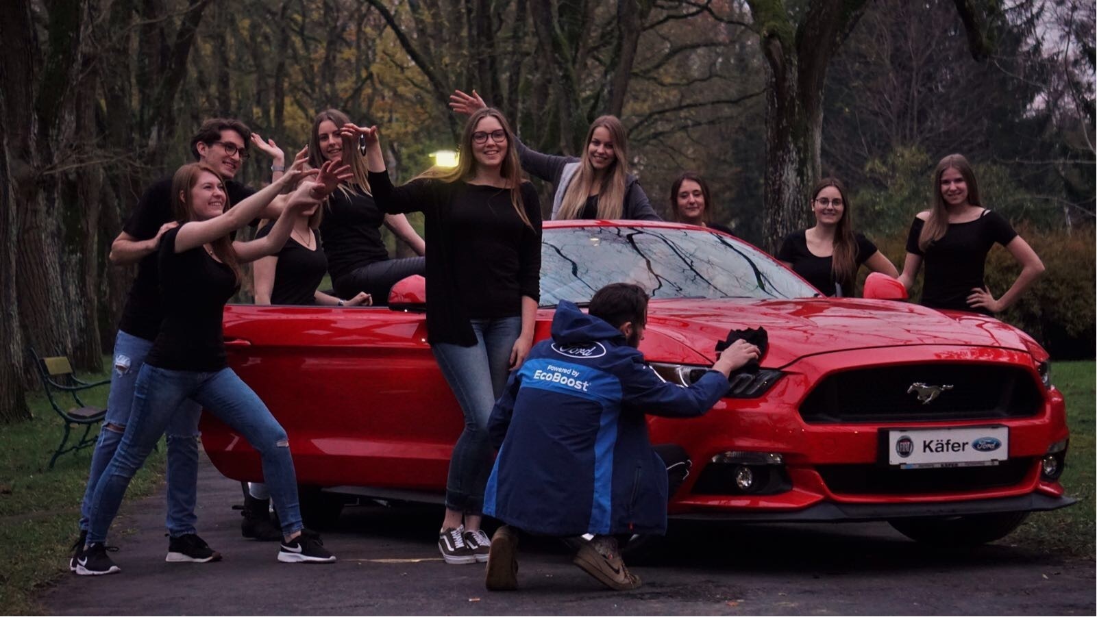 Gruppe junger Leute posiert um ein rotes Ford Mustang Auto in der Natur, während ein Fotograf ein Foto macht.