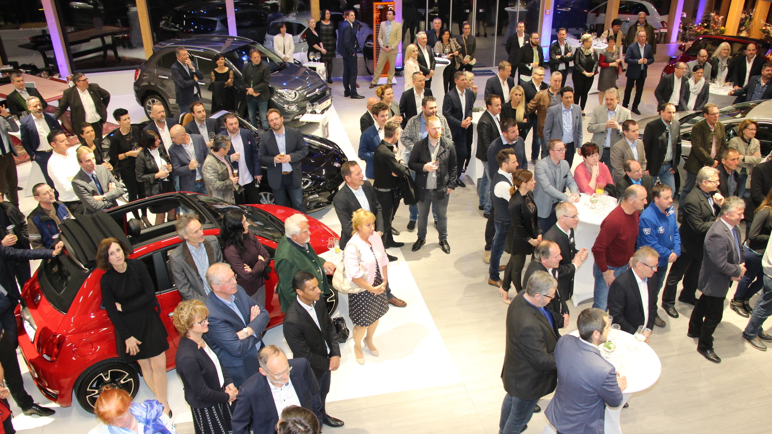 Menschenmenge bei Automobilausstellung in modernem Showroom, verschiedene Fahrzeuge im Hintergrund.