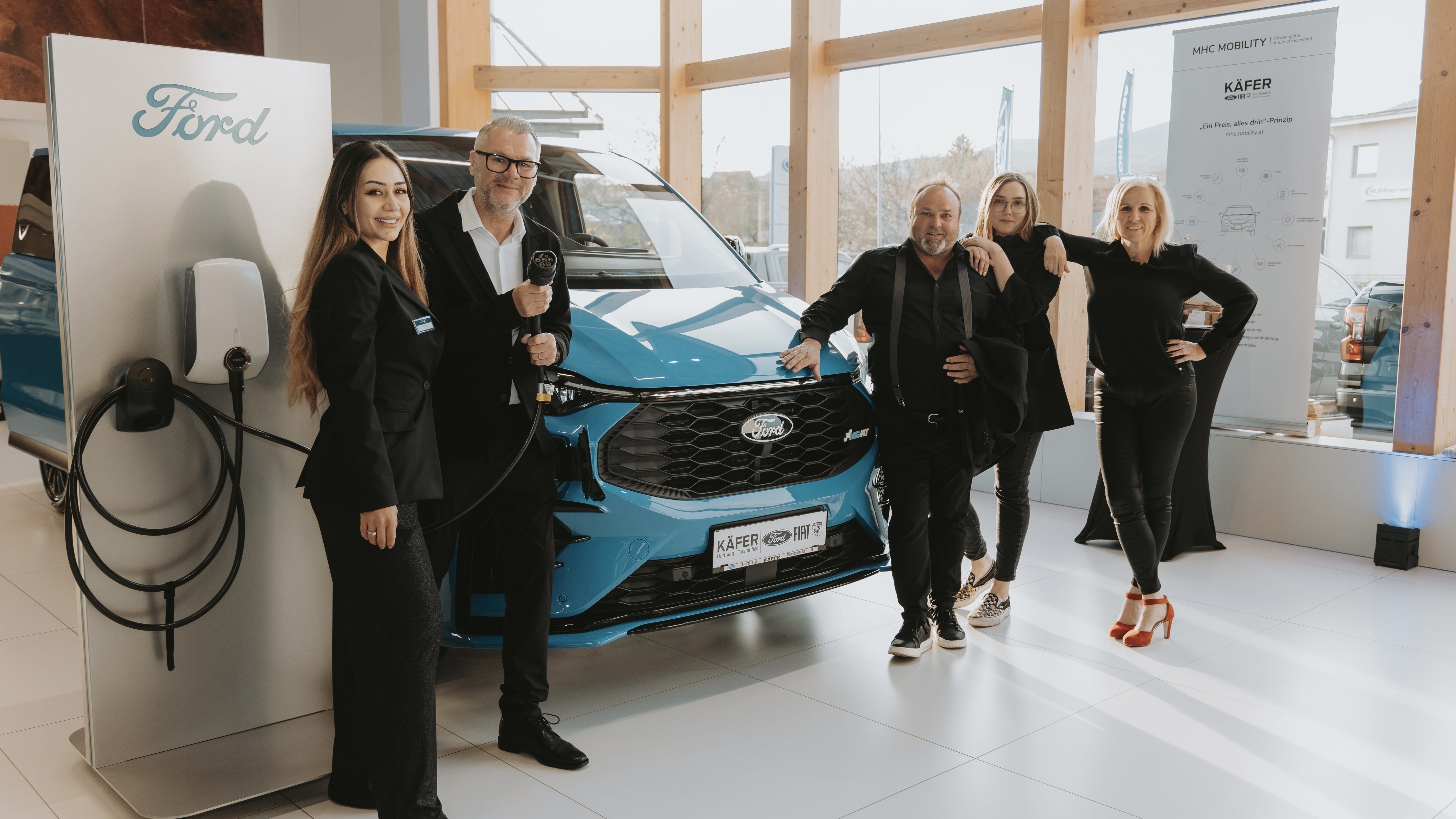 Eine Gruppe von fünf Personen posiert in einem modernen Raum neben einem blauen Ford-Elektrofahrzeug und einer Ladestation.