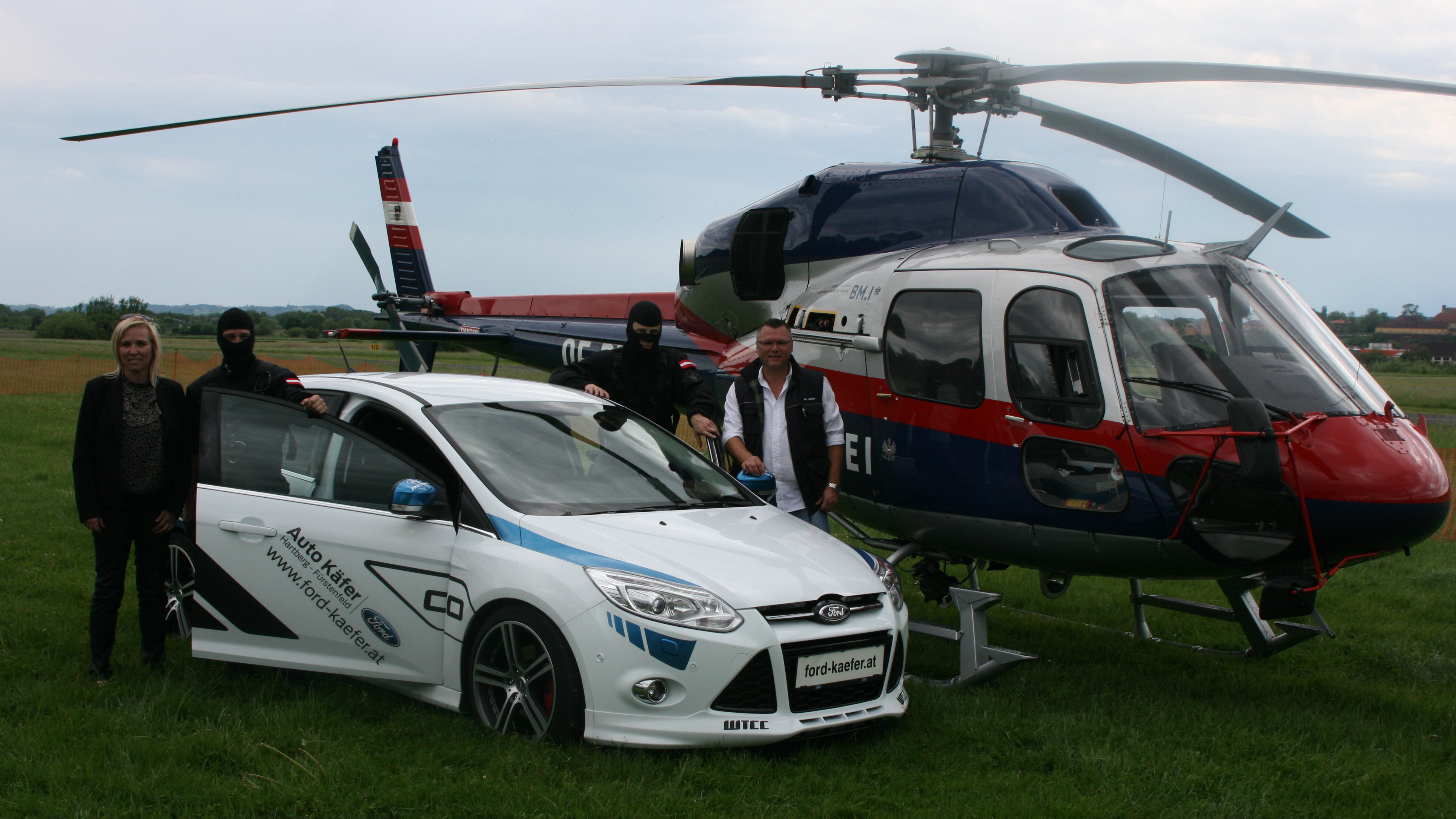 Zwei Fahrzeuge, ein Ford und ein Hubschrauber, stehen auf einem offenen Feld. Drei Personen sind sichtbar.