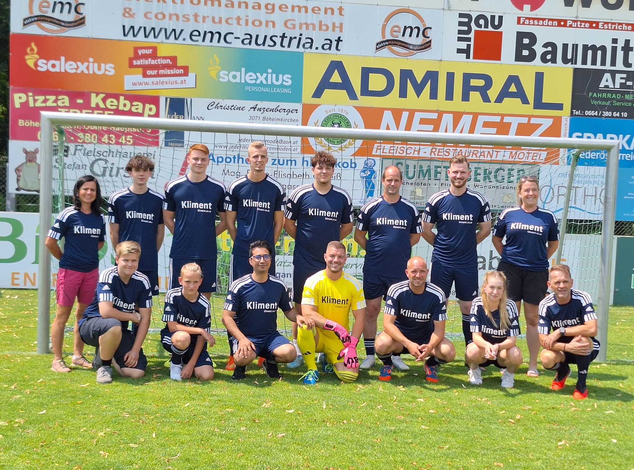 Fußballmannschaft steht vor dem Tor, alle in blauen Trikots mit 'Klinent', mit Sponsorenbanner im Hintergrund.