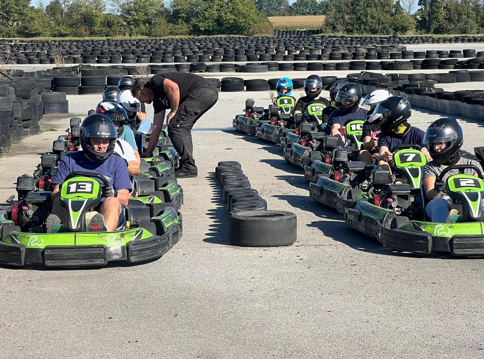 Grupp von Kartfahrern in voller Ausrüstung, bereit auf einer Kartbahn, mit Reifen im Hintergrund.