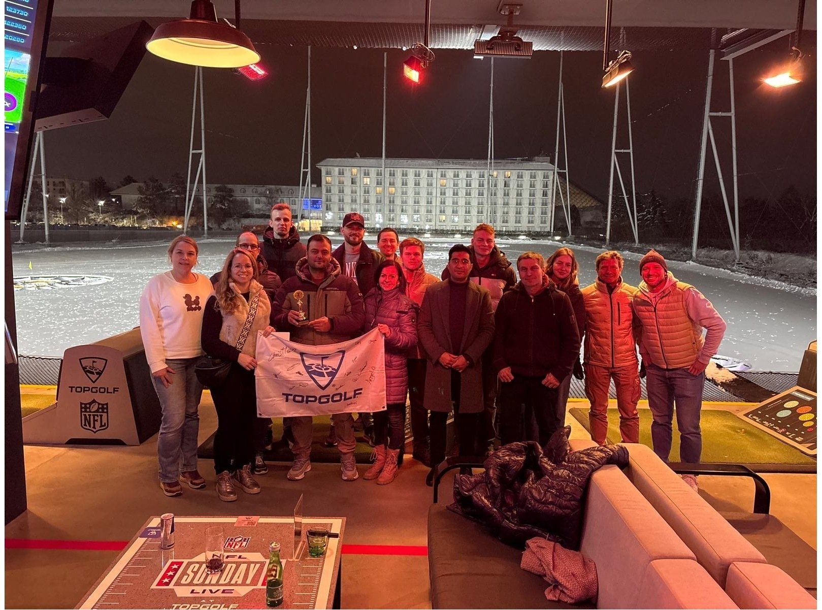 Gruppe von Personen bei Topgolf in der Nacht, hält ein Banner mit dem Topgolf-Logo, winterliche Atmosphäre.